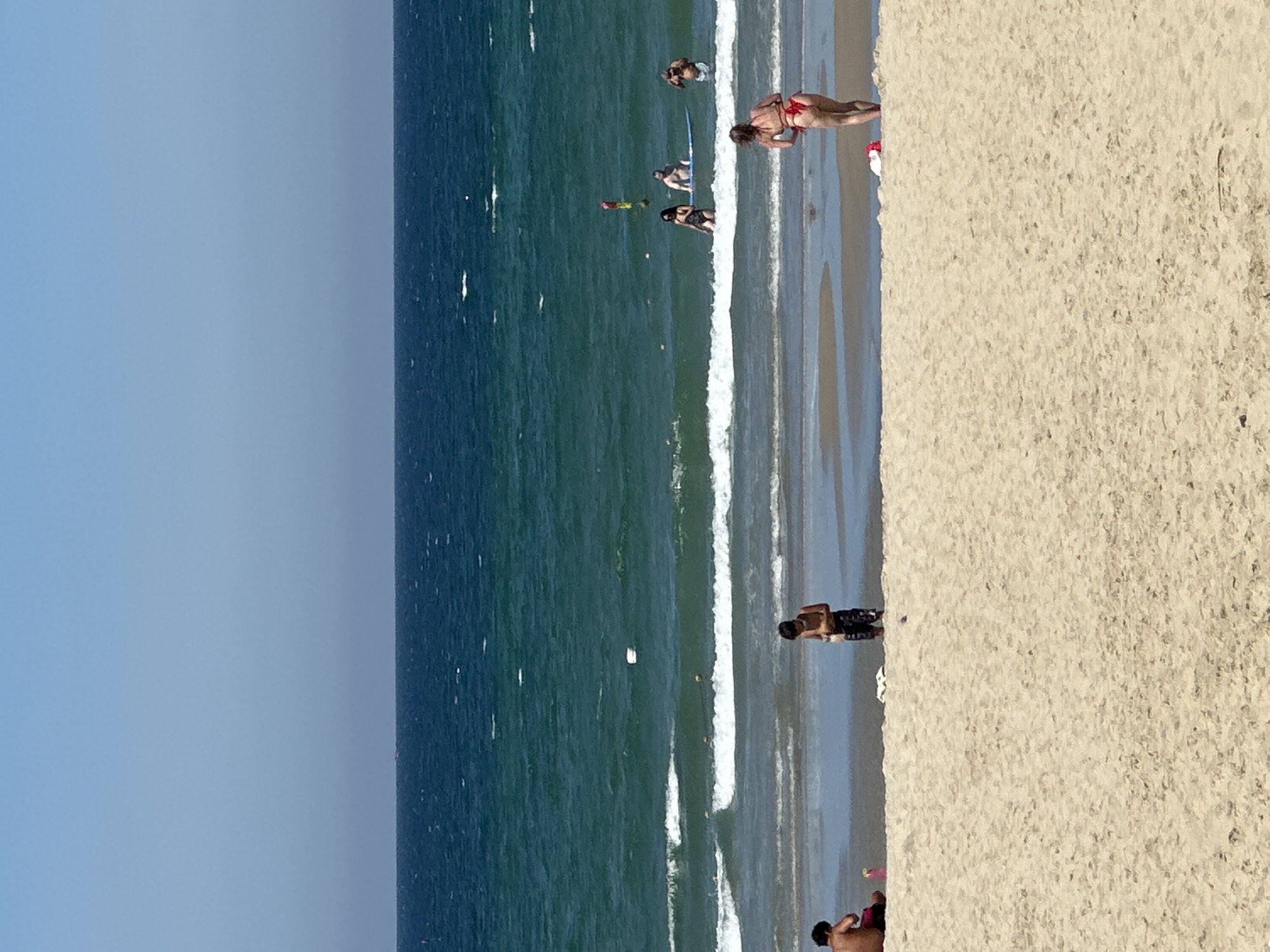 This image captures a sunny day at My Khe Beach in Da Nang, Vietnam, characterized by its wide expanse of golden sand and the vibrant turquoise waters of the East Sea. The scene is divided into three distinct horizontal layers: the sandy beach in the foreground, the breaking waves and shallow water in the middle ground, and the deep blue ocean stretching to the horizon under a clear blue sky.
In the foreground, the sandy beach is largely empty, with the focus on the texture of the sand. On the left, a person is sitting on the sand, facing away from the camera, with a pink bottle nearby. A young boy stands near the water's edge in the middle of the frame, looking down at something in his hands. Further to the right, a woman in a red bikini is facing away from the camera, looking towards the ocean. A white towel and what appears to be a red bag lie on the sand behind her.
The middle ground is dominated by the ocean. Gentle waves are rolling onto the shore, creating white foam. Several people are visible in the water. A group of three individuals are standing in the shallow water further out. Two of them, a man and a woman, appear to be talking. Another man is further out, partially submerged, and another person is on a blue surfboard. A red and yellow buoy with a flag stands out in the water. Small, white buoys or markers are also scattered across the water's surface.
The background features the vast, deep blue ocean extending to the horizon, where it meets the clear sky. A faint, dark line suggesting distant land or ships is visible on the horizon. The overall impression is one of a relaxed beach day with clear weather and moderate activity in the water.