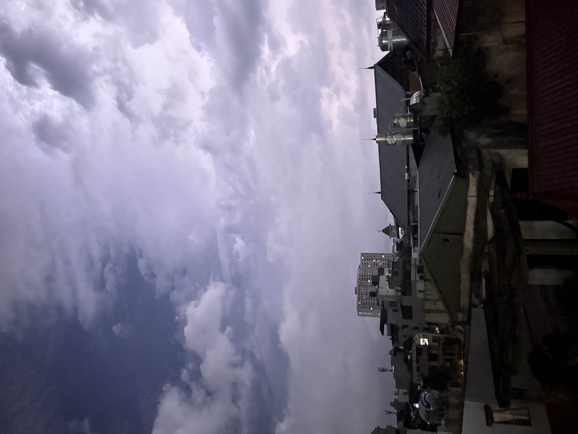 The image shows a nighttime urban scene in Hà Nội, Vietnam, with a dramatic sky filled with dark, stormy clouds. Lightning flashes are visible in the clouds. The foreground features rooftops of buildings, some with antennas and water tanks. In the distance, there is a high-rise building with lighted windows. The lighting suggests a thunderstorm is in progress. No people are visible.