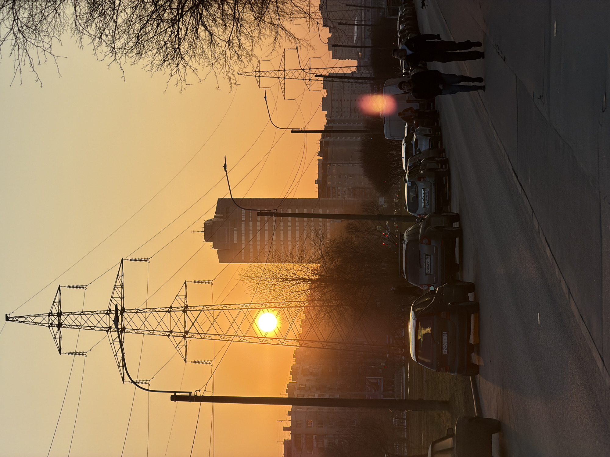 The image captures a sunset scene in Saint Petersburg, Russia. The sun is positioned behind an electrical pylon, creating a silhouette effect. In the background, there are several buildings and trees. A row of parked cars lines the side of the road, and a couple of people are walking on the sidewalk alongside the cars. The sky is a gradient of orange and yellow, indicating the time of day.