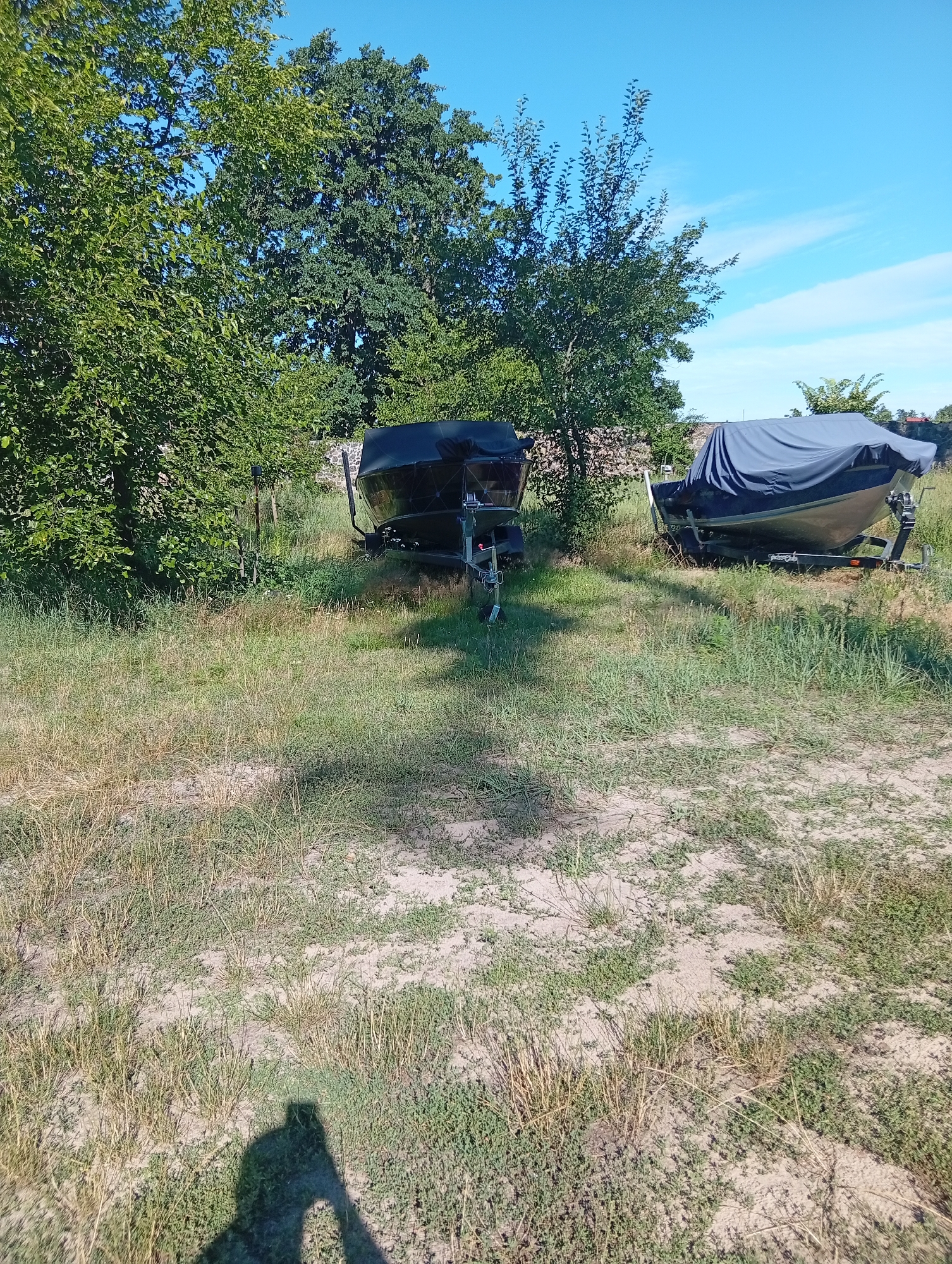 The image depicts an outdoor scene in Sokyrna, Ukraine, with two boats on trailers parked in a grassy area surrounded by trees under a clear blue sky. The boat on the left is dark and uncovered, with a trailer visible. The boat on the right is covered with a dark grey tarp. The ground is covered with dry grass and patches of bare soil. The foreground shows the shadow of a person taking the photograph. The environment suggests a storage or recreational area, possibly near a body of water, given the presence of boats.