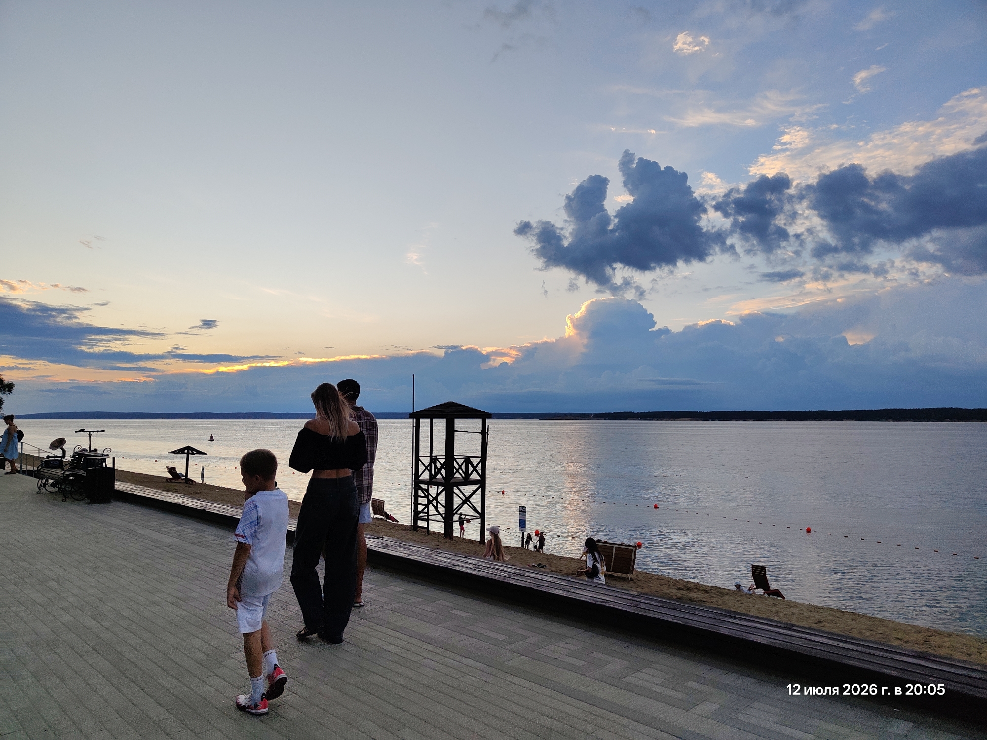 The image shows a waterfront scene at dusk, likely a beach or promenade. In the foreground, a young boy in a white shirt and shorts walks away from the camera. To his right, a woman in a black crop top and dark pants stands with her back to the camera, her arm around a man in a plaid shirt and shorts. The promenade is paved with light-colored rectangular tiles.
Beyond the promenade is a sandy beach with several people. A few are sitting or playing near the water's edge, and a lifeguard tower stands prominently on the sand. Red buoys mark a swimming area in the calm water. The lake stretches out to the horizon, where a tree line is visible under a dramatic sky filled with clouds illuminated by the setting sun. The timestamp in the bottom right corner reads "12 июля 2026 г. в 20:05".