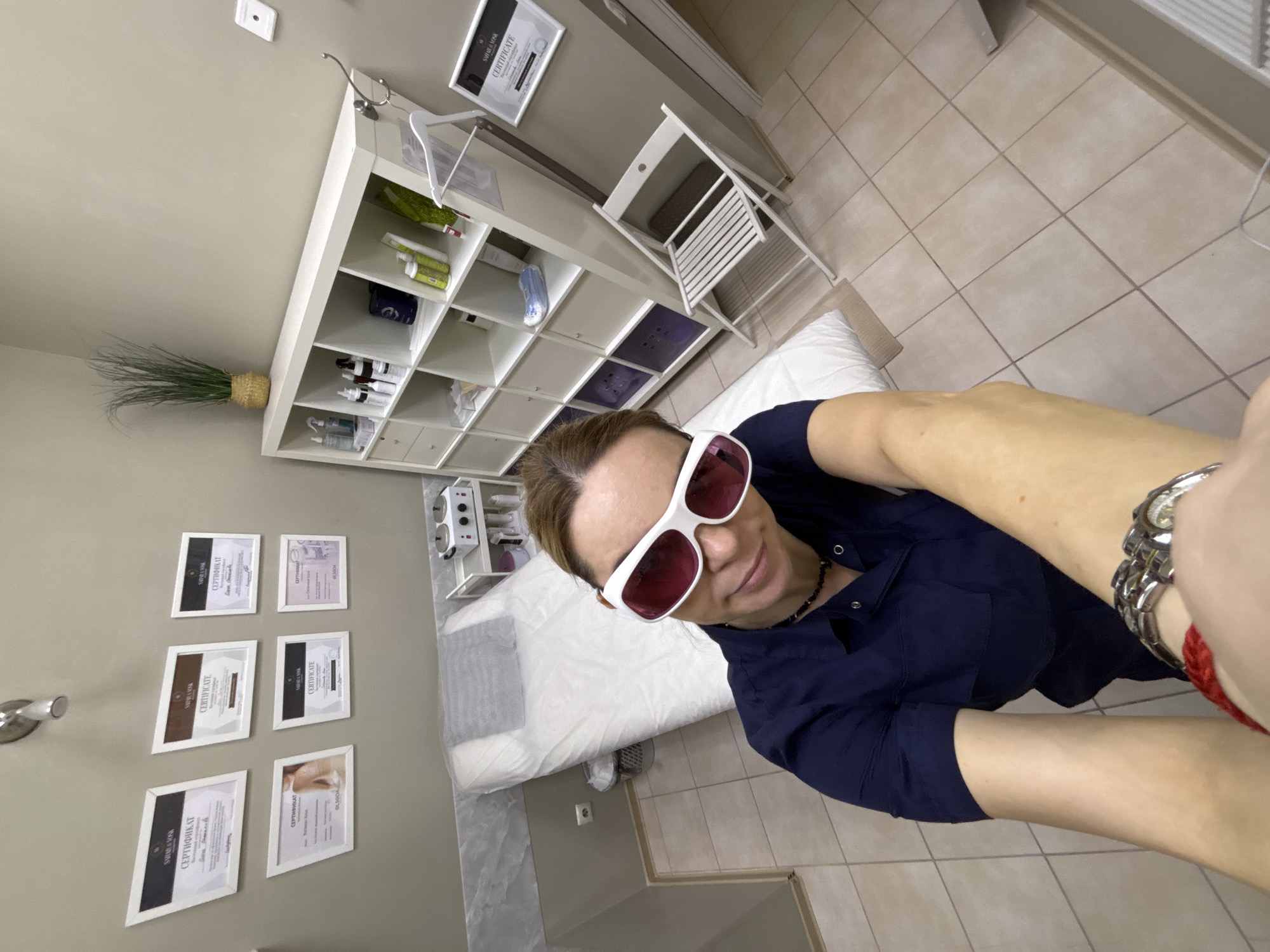 The image appears to be a selfie taken inside what seems to be a beauty salon or medical office. The person taking the photo is wearing white sunglasses with red lenses and a dark blue collared shirt. In the background, there's a medical bed with white linen, a white shelving unit holding various products and supplies, and certificates displayed on the wall. A folding chair is also visible. The floor is tiled. There is no information available about the location in Vsevolozhsk, Russia, but based on the interior design, it could be any business related to beauty treatments or medical aesthetics.