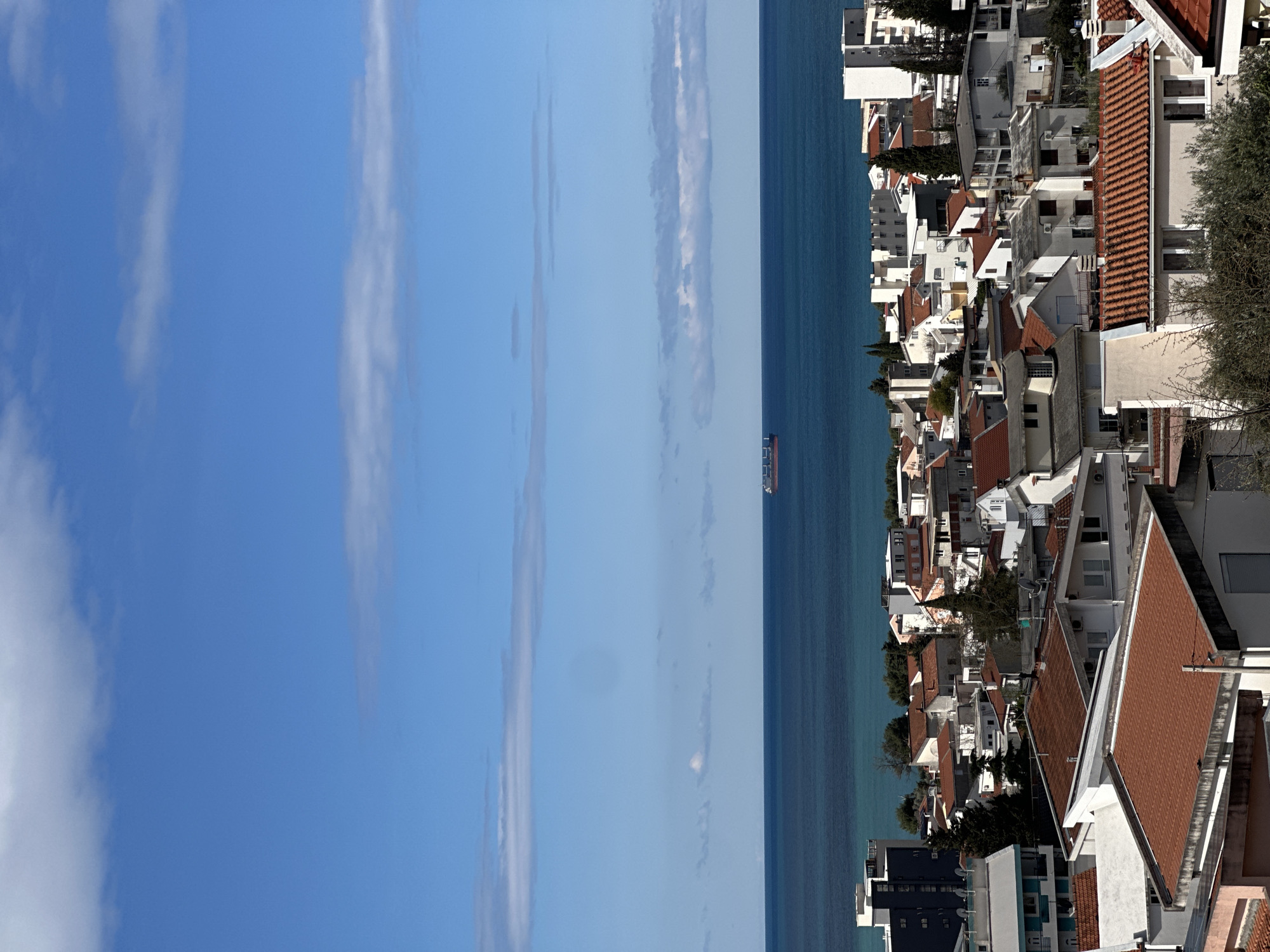 The image shows a view of the coast of Šušanj, Montenegro. The sky is blue with thin, long clouds stretching vertically. The Adriatic Sea is a deep blue, with a cargo ship visible in the distance. On the right side of the image, there is a densely populated area consisting of buildings and houses, mostly white with red-tiled roofs. The buildings appear to be situated on a hillside, descending towards the sea. There is some sparse greenery among the buildings.