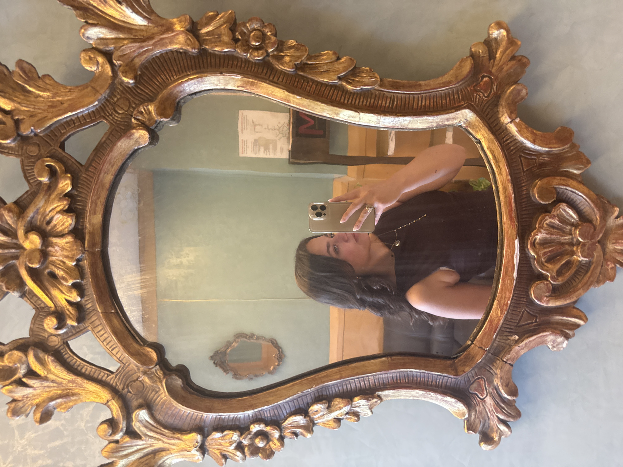 A woman is taking a selfie in a gold-framed ornate mirror. The mirror is reflecting the room, including another, smaller ornate mirror on the wall. The room has light blue walls and wooden trim. There is a diagram or sign on the wall behind the woman, and boxes or furniture are visible in the background. Based on visual characteristics, it's not possible to determine if the location is in Sofia, Bulgaria.
