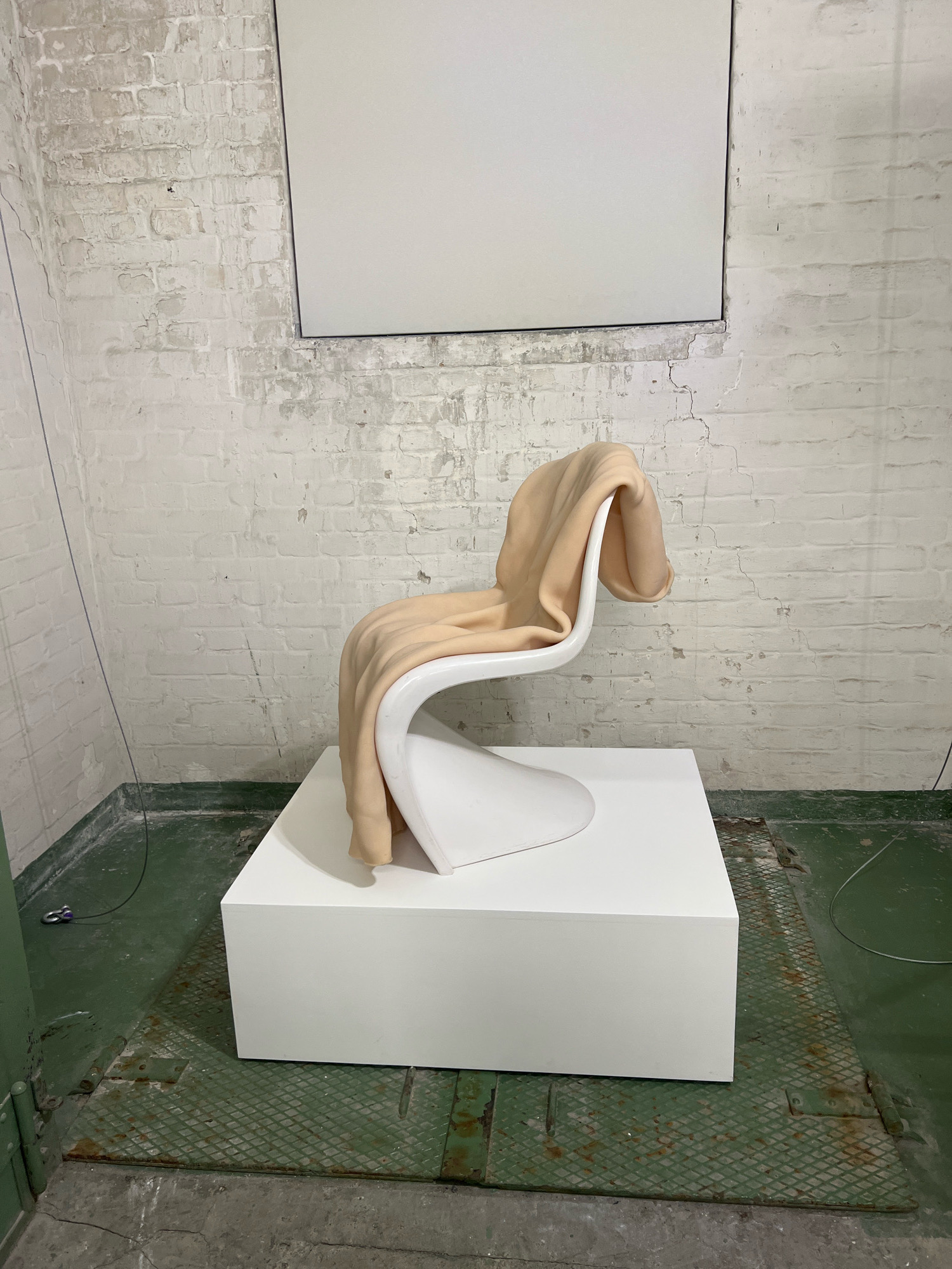The image depicts an indoor scene, likely in a studio or exhibition space, with a focus on a white pedestal that supports a distinctive, curved white chair. A light peach-colored blanket is draped over the chair. The background consists of a white brick wall, showing signs of age and wear, with a large, plain rectangular panel mounted high on the wall. The floor is a mix of concrete and green-painted metal grating, with some electrical cords visible. The overall aesthetic suggests an industrial or minimalist setting. There is no discernible indication of the location being in Berlin, Germany. No people are present in the image.