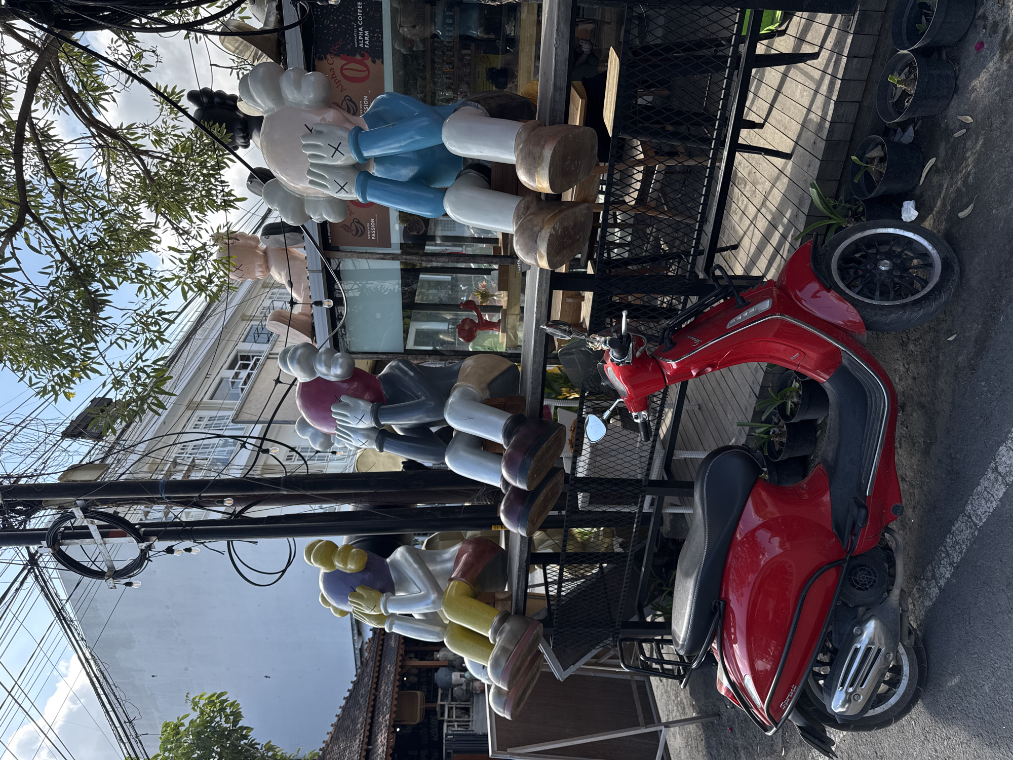 In North Kuta, Indonesia, a bright red scooter is parked on a street in front of a cafe with outdoor seating. Several large, cartoon-like figures with their hands covering their eyes are seated on a wooden bench in front of the cafe. The figures are colorful and appear to be made of plastic or fiberglass. Behind the figures and the cafe, there are buildings, trees, and a sky with some clouds. A complex network of electrical wires hangs overhead.