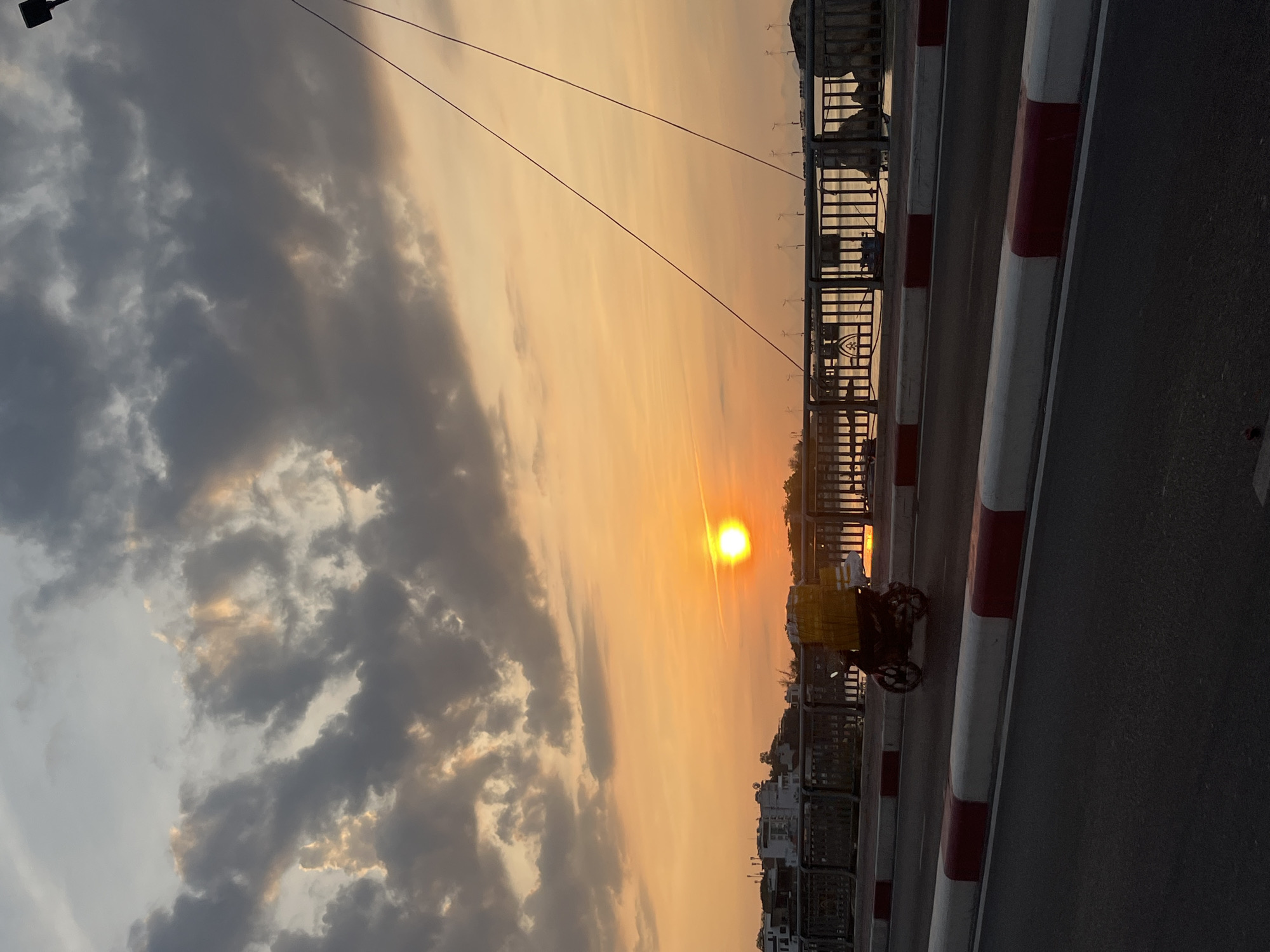 The image captures a sunset in Bac Nha Trang Ward, Vietnam. The sun is setting directly in front of the viewpoint, casting an orange glow across the sky and clouds. A motorcycle with a large yellow bag is parked on the right side of the road. The road is bordered by a red and white striped barrier. Beyond the barrier, there is a metal railing overlooking the water and buildings. The background features buildings silhouetted against the setting sun. Utility lines are also visible overhead. The sky has a mix of light orange hues and dark, cloudy patches. The viewpoint is most likely on the Tran Phu Bridge.