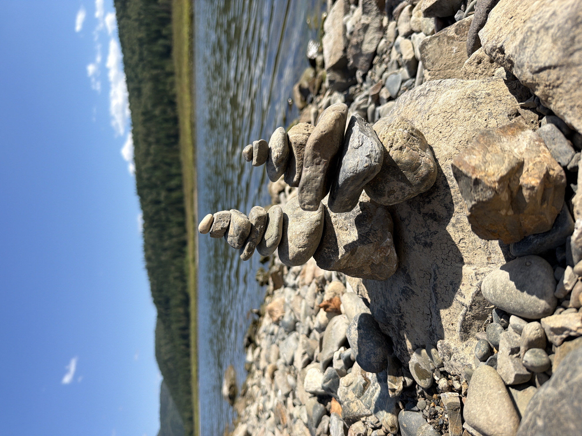 This is a close-up, low-angle outdoor photograph of two cairns made of stacked river stones on a rocky shore. The cairns are the primary subjects, positioned in the foreground of the image. The left cairn is taller and more slender, composed of approximately seven stones of varying sizes and shades of grey and tan. The right cairn is shorter and wider, with about five stones.
The shore is covered in a multitude of similar river stones, ranging from small pebbles to larger, fist-sized rocks. Some of these stones are partially submerged in the shallow water.
In the background, a body of water, likely a river or lake, stretches across the mid-ground, its surface rippled by the breeze and reflecting the sunlight. Beyond the water, a densely forested hillside rises, its green foliage contrasting with the blue sky. A few wispy white clouds are visible in the upper portion of the sky. The overall environment suggests a natural, serene setting, possibly in or near the village of Ust-Kabirza, Russia, a location known for its proximity to the Kuznetsk Alatau Nature Reserve and its numerous rivers and forests.
There are no people or animals visible in the image. The lighting indicates it is daytime, with the sun casting distinct shadows on the stones, particularly on the larger rock supporting the left cairn. The focus is sharp on the cairns and the immediate rocky foreground, with the background elements becoming progressively softer and blurred, creating a sense of depth.