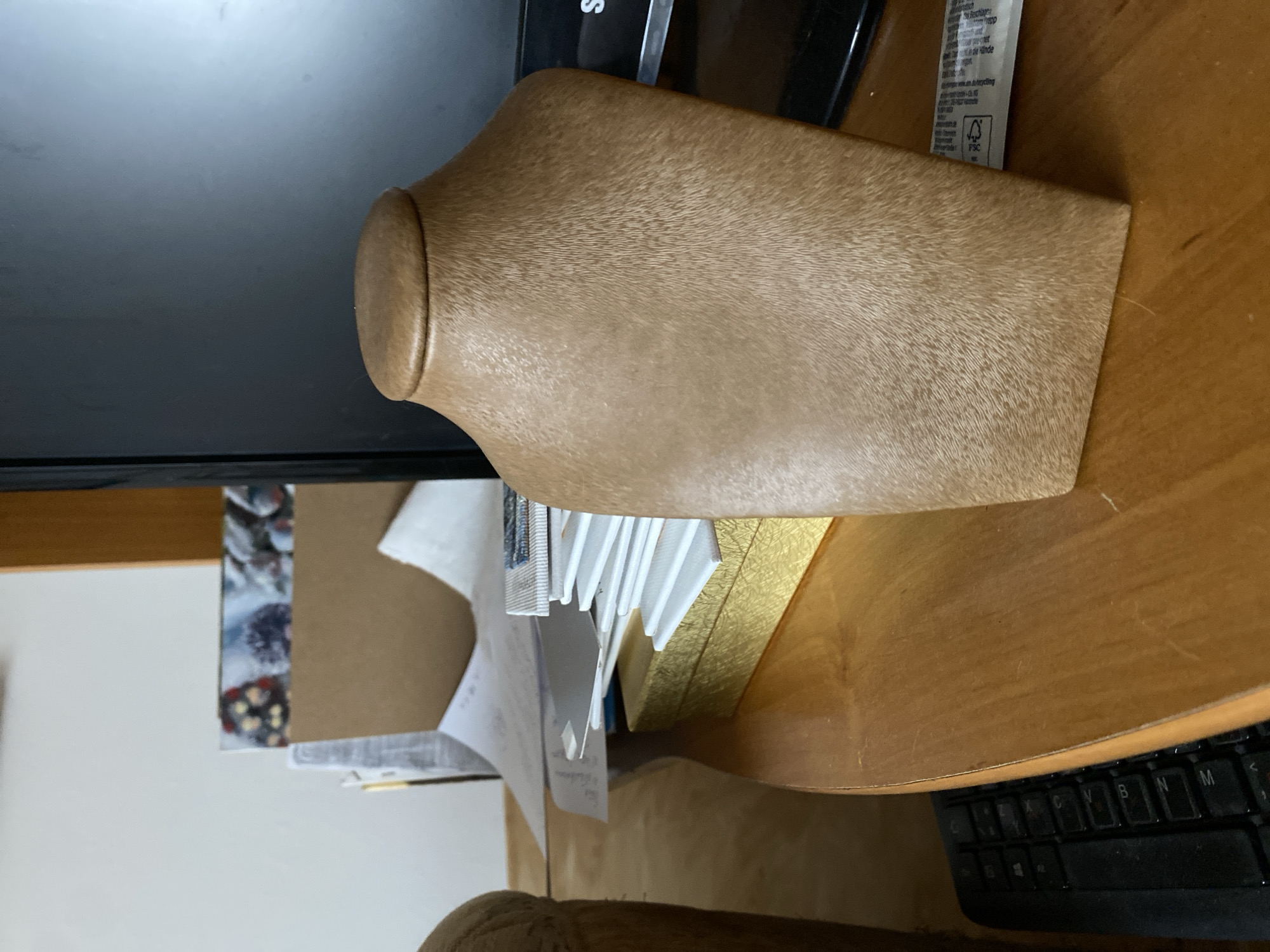 The image shows a close-up view of a brown necklace bust placed on a wooden desk. The bust is positioned in the foreground and is the primary subject of the photograph. To the left of the bust, a black keyboard is partially visible, suggesting a workspace. In the background, there are stacks of paper, a golden box, and a monitor. The overall scene appears to be an indoor setting, likely an office or home study. The lighting is moderate, casting soft shadows. The image does not contain any information that would allow for a specific location in Bílina, Czechia to be identified.
