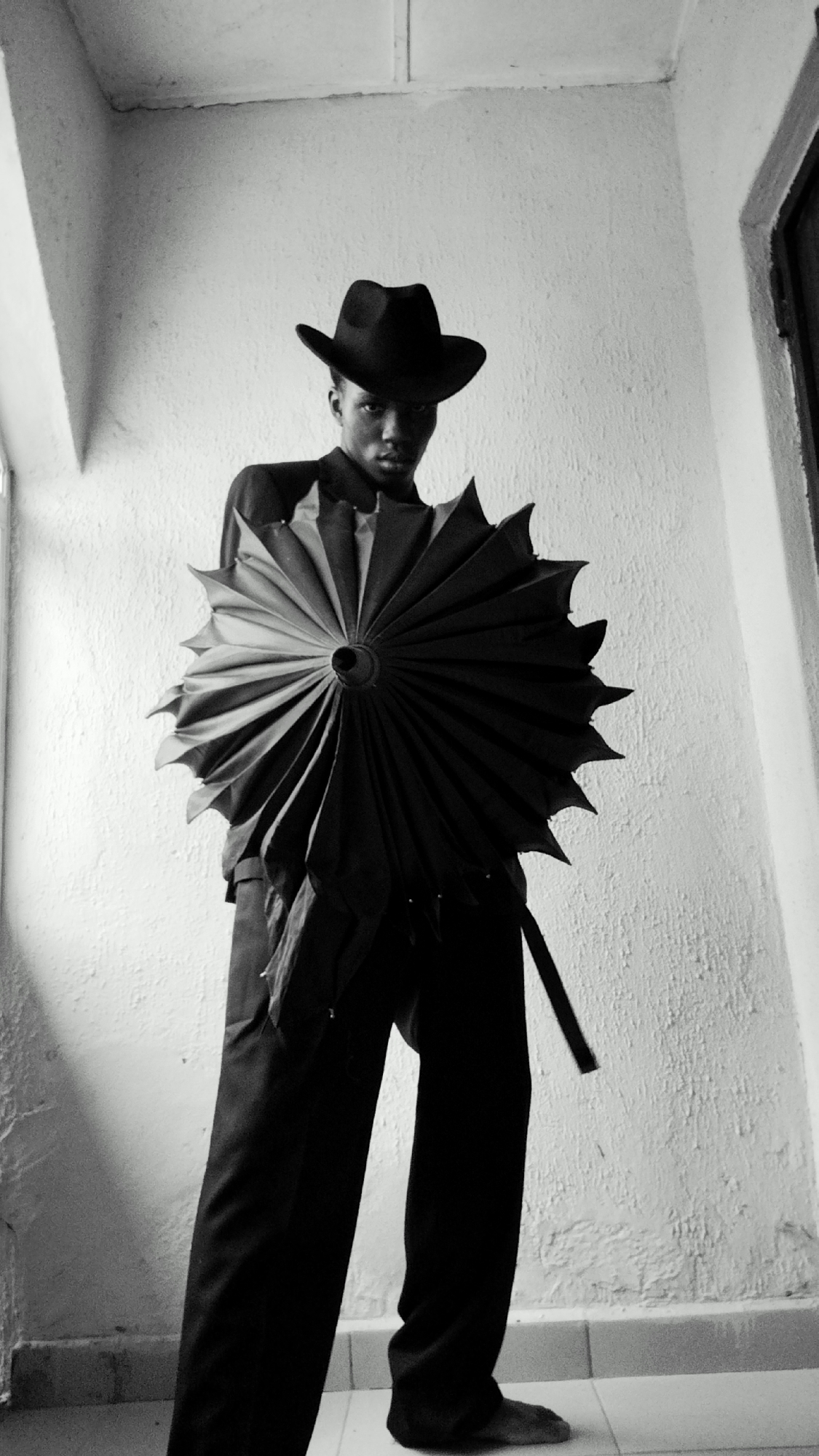 A full-body, low-angle, black and white photograph shows a young Black male standing indoors, holding a large, black, intricately folded umbrella open in front of himself. He is wearing a dark fedora hat, a collared shirt under a dark jacket, and dark trousers. His face is turned towards the camera, and he is looking directly at the viewer. The umbrella is positioned centrally in the frame, its spokes creating a starburst pattern. The background consists of a textured white wall and a tiled floor. The lighting is stark, casting shadows that emphasize the textures of the wall and the folds of the umbrella. The scene appears to be an interior space, possibly a hallway or a room with minimal furnishings, located in Sabon Tasha, Nigeria.