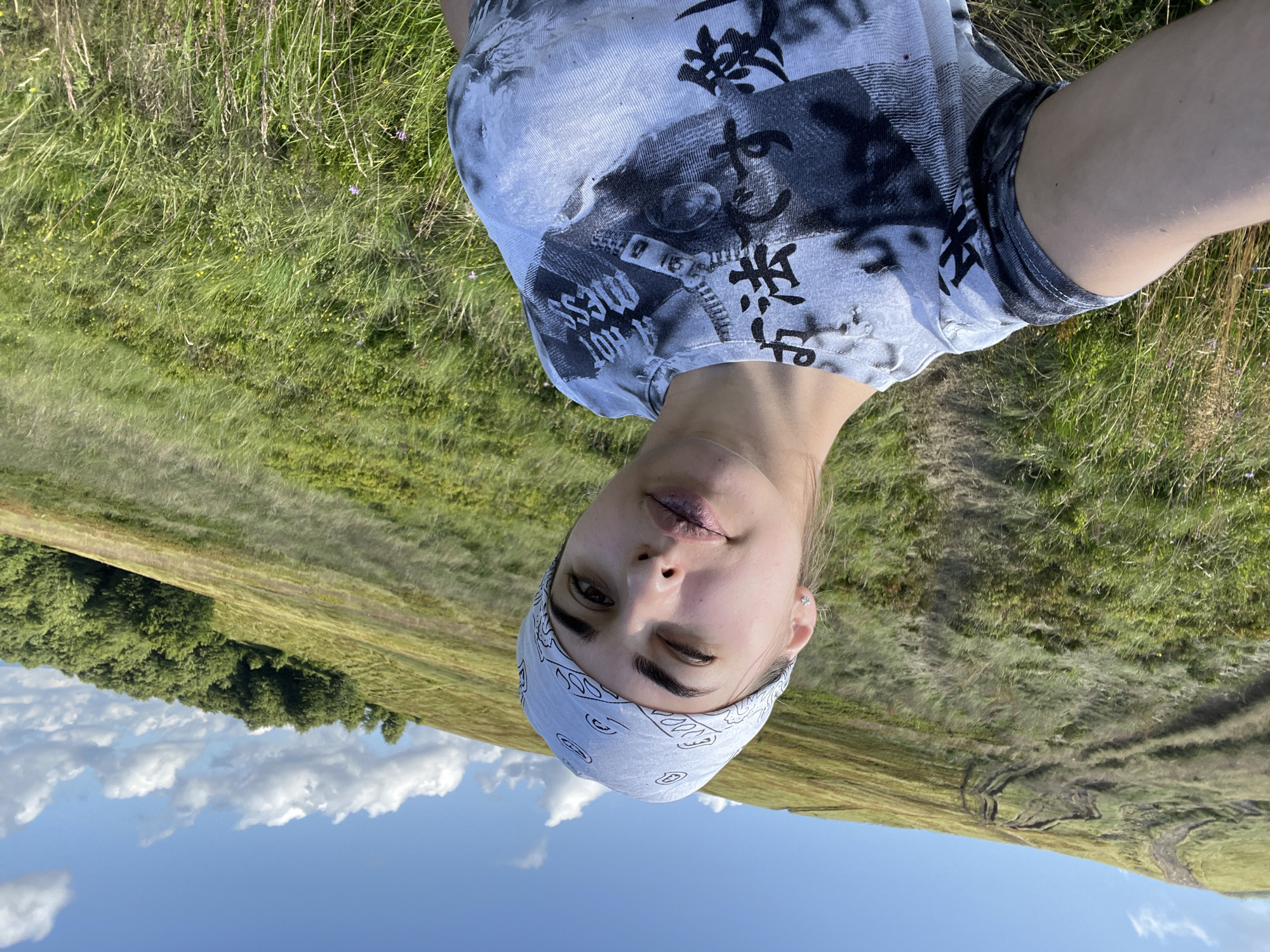 A young woman, wearing a light blue bandana with a white paisley pattern and a grey t-shirt with a black graphic print, takes a selfie on a grassy hillside. The woman is looking towards the camera, with her left eye partially closed. Her lips are parted, and she appears to be mid-speech or in the process of blinking. The background consists of a rolling green hillside with a dirt track winding up it, and a distant treeline on the right side of the frame. The sky above is blue with scattered white clouds. The image appears to have been taken outdoors, in a natural, possibly mountainous, environment, likely in the vicinity of Ust'-Chorna, Ukraine, given the landscape features.