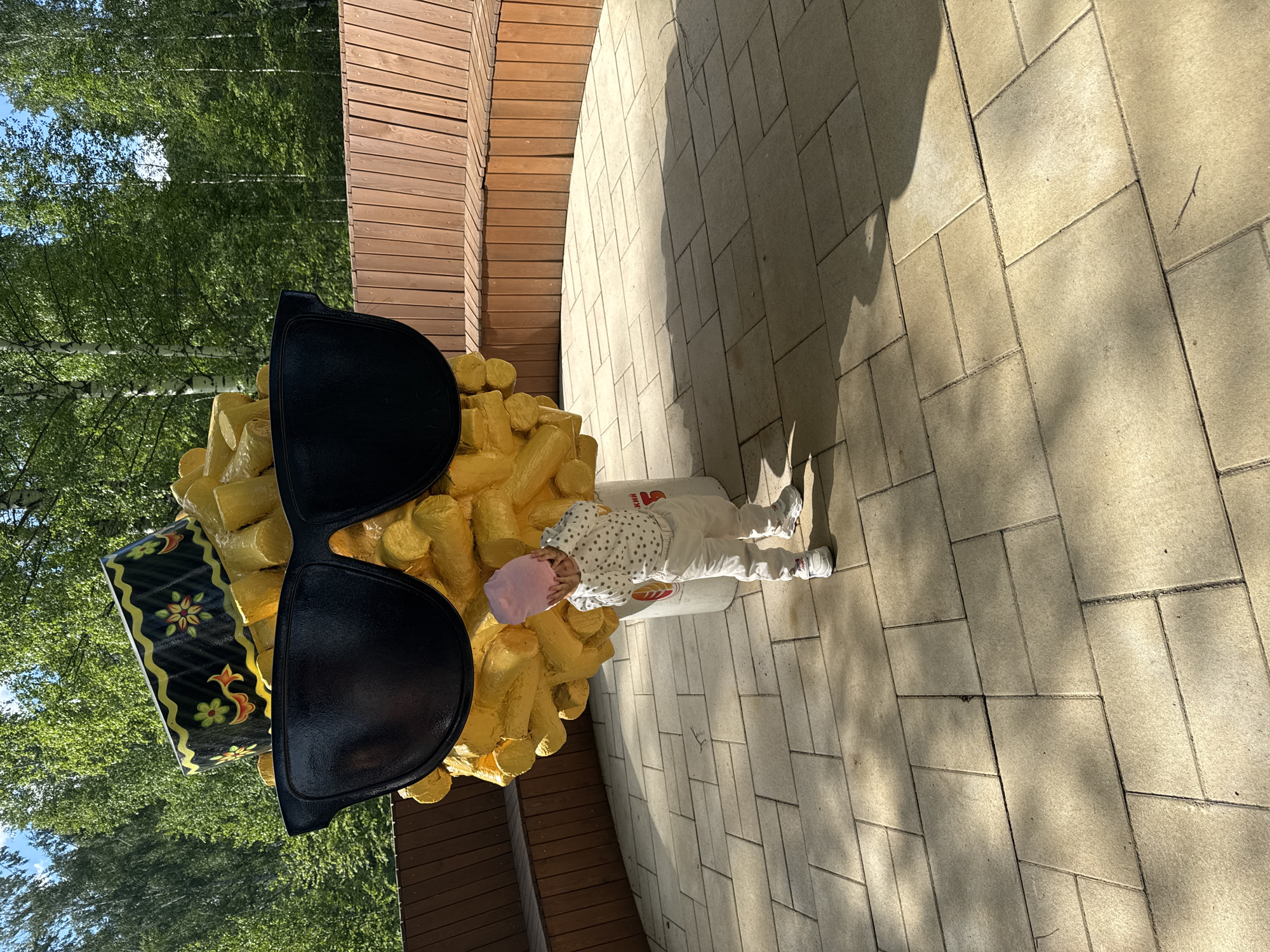 A child in a pink cap and white clothing is standing next to a large sculpture of french fries with sunglasses in Zelenodolsk, Russia. The sculpture is painted yellow and is next to a wooden wall. The ground is paved with rectangular stones. Trees are visible in the background.