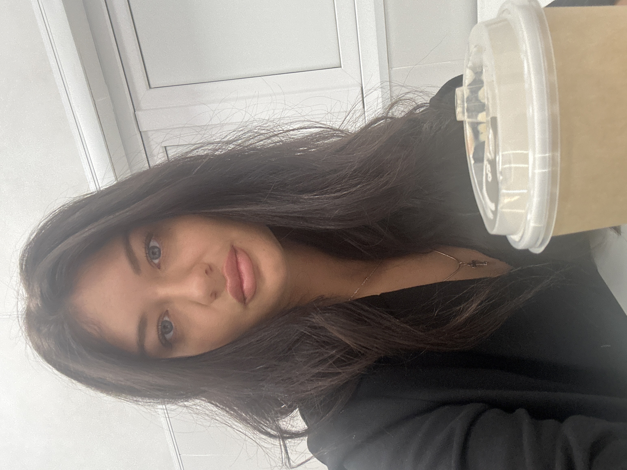 A close-up, slightly rotated portrait-style image features a young woman with dark hair, looking directly towards the viewer. Her face is in the left half of the frame, with her dark, long, and slightly wavy hair framing her face and extending across her shoulders. She has fair skin, full pink lips, and visible light-colored eyes. She appears to be wearing a dark top, possibly black, and a delicate gold-toned necklace with a small pendant is visible around her neck.
To the right side of the frame, a disposable light brown or beige paper cup with a clear plastic lid is prominently featured, held or positioned near her. The contents of the cup are not clearly visible but appear to be a light-colored beverage. The background is a bright, light-colored (likely white or off-white) wall or ceiling, with faint dark lines suggesting architectural details such as window frames or ceiling panels.
The lighting is bright and even, indicating an indoor setting, possibly during the daytime. The woman's expression is neutral, looking towards the camera. No discernible text is clearly readable on the cup lid or in the background.