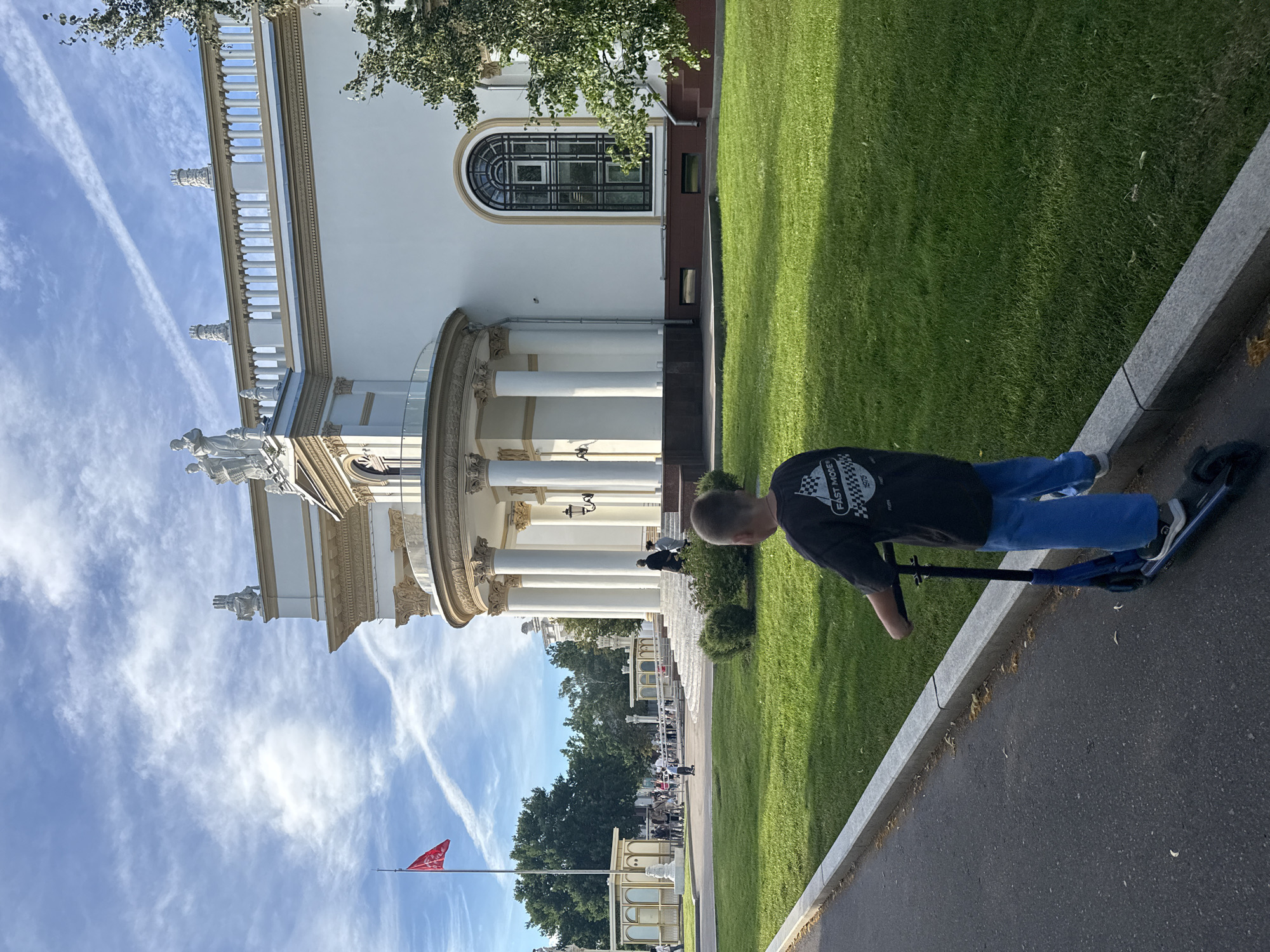 The image depicts a young boy riding a blue scooter on an asphalt path bordering a manicured green lawn. He is wearing a black t-shirt with a checkered graphic and blue pants. In the background, a large, ornate white building with classical architectural elements, including columns, a rounded portico, and decorative statues, stands under a partly cloudy blue sky. A red flag is visible on a flagpole to the left of the building. Several trees and smaller structures are also present on the grounds, and a few other people are seen in the distance, some walking and others near the building's entrance. The scene appears to be a public park or an exhibition ground. The architectural style and the presence of a Soviet-era flag suggest the location could be VDNKh (Exhibition of Achievements of National Economy) in Moscow, Russia, known for its grand pavilions and open spaces.