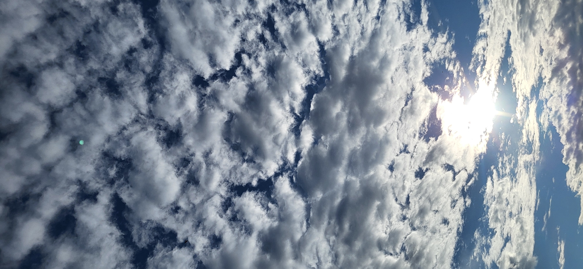 This image captures a dynamic sky filled with a mixture of fluffy white clouds and darker, heavier cloud formations against a vibrant blue sky. The sun is visible in the upper right quadrant of the image, shining brightly and creating a lens flare effect. The clouds appear to be moving or forming, suggesting a day with changing weather conditions. The orientation of the image is vertical, with the sun appearing on the right side. There is a small, circular, bright turquoise object visible in the sky, positioned to the left of the center. This could be a photographic artifact or a distant object. Another small dark object, possibly a bird or insect, is visible in the upper left portion of the frame. The scene evokes a sense of openness and the vastness of the sky. The lighting suggests it is likely daytime, with the sun's position indicating it could be morning or afternoon. No text is visible in the image. The location context provided is Hetmanivka, Ukraine.