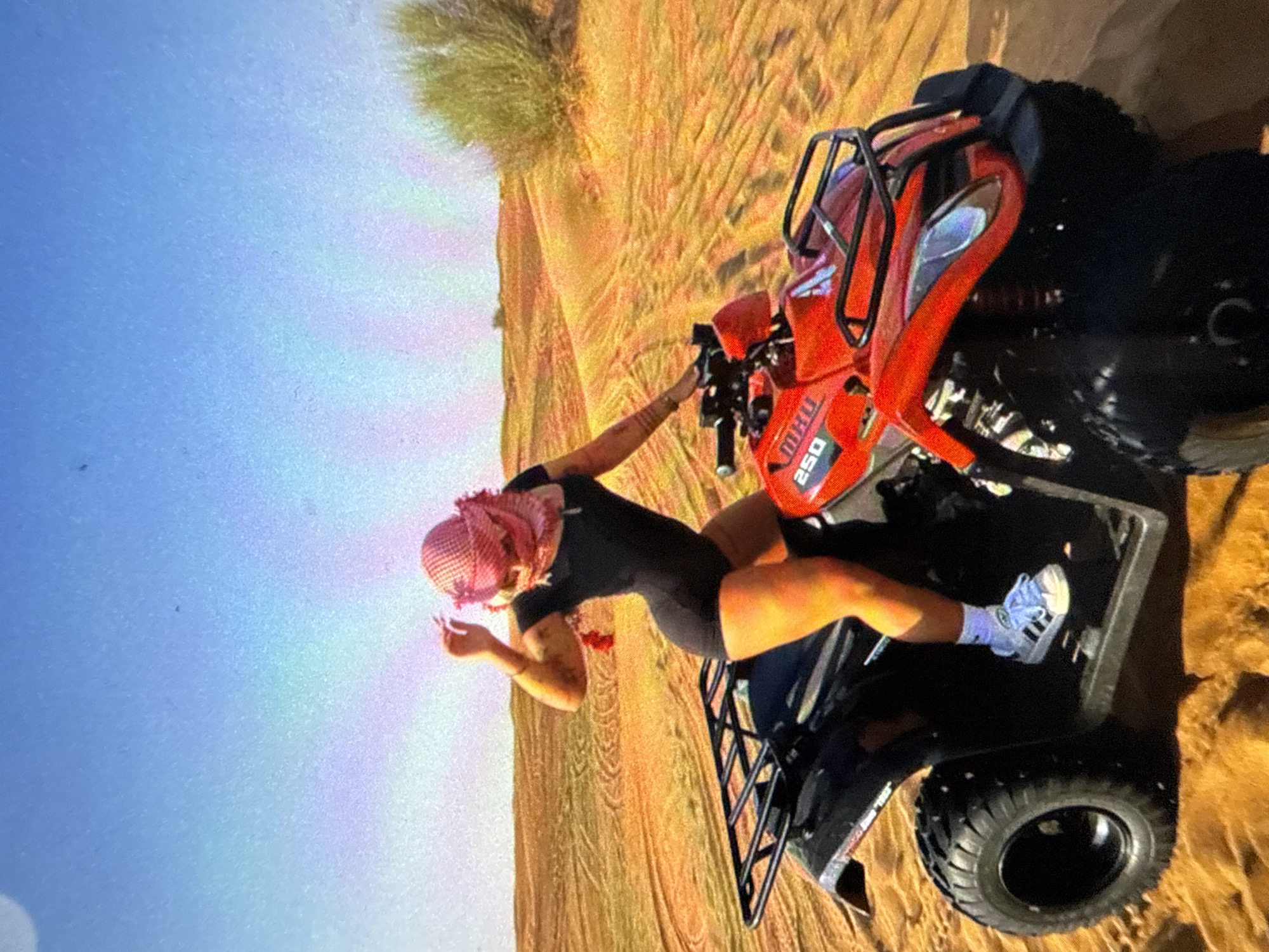 The image depicts a person in a black outfit and a red-and-white head covering posing with a red ATV on a sand dune. The setting appears to be a desert landscape under a blue sky. The ATV has "250" displayed on its side. The location is Unknown, Unknown.