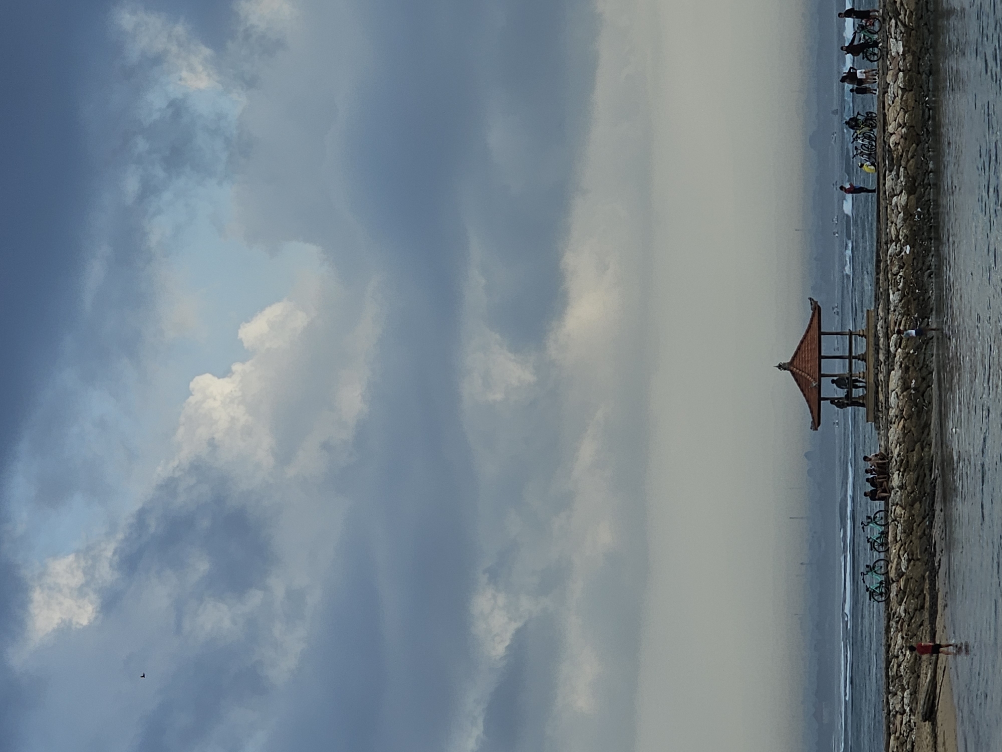 A wide shot captures a section of the coastline in Denpasar, Indonesia, under a dramatic, cloudy sky. In the foreground, calm water laps against a stone jetty. Several people are visible on the jetty, some standing and conversing, while others are seated. Three bicycles are parked near a small group. Further along the jetty, a traditional Indonesian gazebo with a red, multi-tiered roof stands prominently. More people are gathered around and inside the gazebo. In the middle ground, the ocean features gentle waves, and a hazy horizon suggests the presence of distant land or buildings. The sky is filled with a mix of dark gray and white clouds, with a small bird soaring in the upper left quadrant. The overall atmosphere is serene, with the natural elements of water and sky dominating the scene.