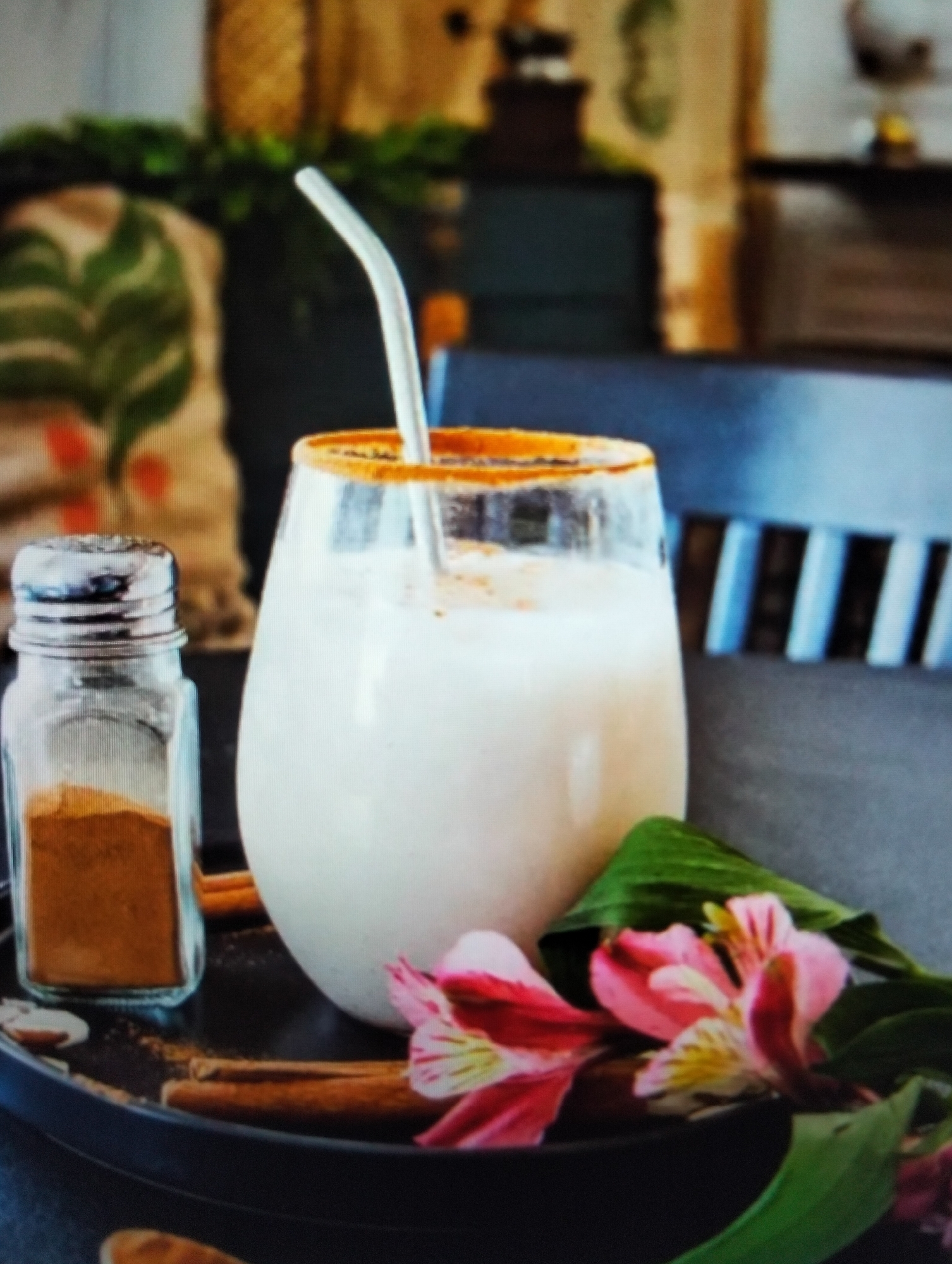 The image shows a close-up view of a glass of creamy white beverage, likely a smoothie or milkshake, with a cinnamon-dusted rim. A metal straw extends from the glass. Next to the glass is a small shaker filled with cinnamon powder. Several cinnamon sticks are scattered on a dark tray in front of the beverage, along with a few pink and yellow flowers and green leaves. The background is blurred, but appears to be an indoor setting with decorative elements like a woven tapestry and dark furniture. The scene is set on a dark, circular tray. The image does not provide any information or visual cues that would allow for the identification of a location in Payagaboh, Indonesia.