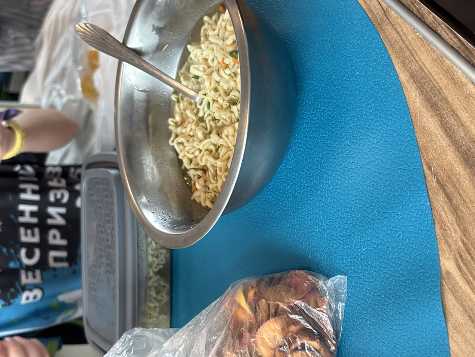 The scene, located in Ivanovka, Russia, depicts an indoor setting focused on a table surface. A stainless steel bowl, exhibiting condensation on its exterior, is positioned prominently on a bright blue, textured placemat. Inside the bowl, prepared instant noodles with visible green and orange dehydrated vegetable fragments are immersed in broth, and a silver spoon with a fluted handle rests within. To the left of the bowl, a transparent plastic bag contains numerous dried, reddish-brown, and yellow fruit or vegetable pieces. Behind these items, a grey rectangular plastic container with a patterned, semi-transparent lid is also visible, containing what appears to be instant noodles. The blue placemat rests on a wooden-patterned table. In the upper background, the upper torso and arms of an individual are partially visible. The person is wearing a dark blue T-shirt featuring prominent white Cyrillic text that reads "ВЕСЕННИЙ ПРИЗЫВ" (VESENNIY PRIZYV), translating to "Spring Conscription" or "Spring Draft." A yellow wristband is present on the individual's right wrist. The background beyond the person is indistinct, appearing as a blurred white fabric.