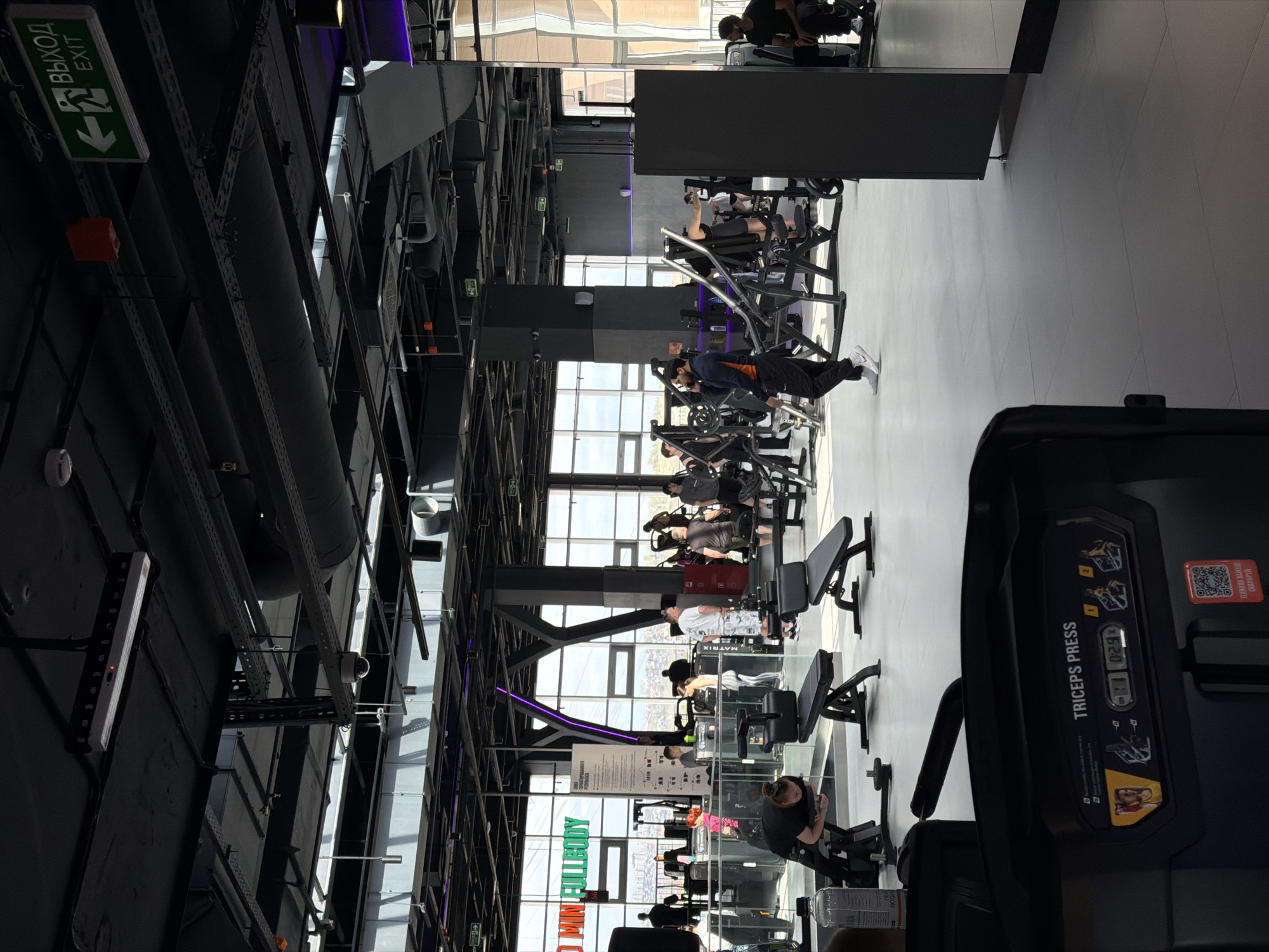 The image depicts the interior of a gym located in Krasnoyarsk, Russia. Several individuals are engaged in exercising on various machines. The gym features a high ceiling with exposed pipes and beams. Large windows provide natural light. Exercise equipment includes triceps press, and other weight machines. An exit sign in Russian and English is visible on the ceiling.