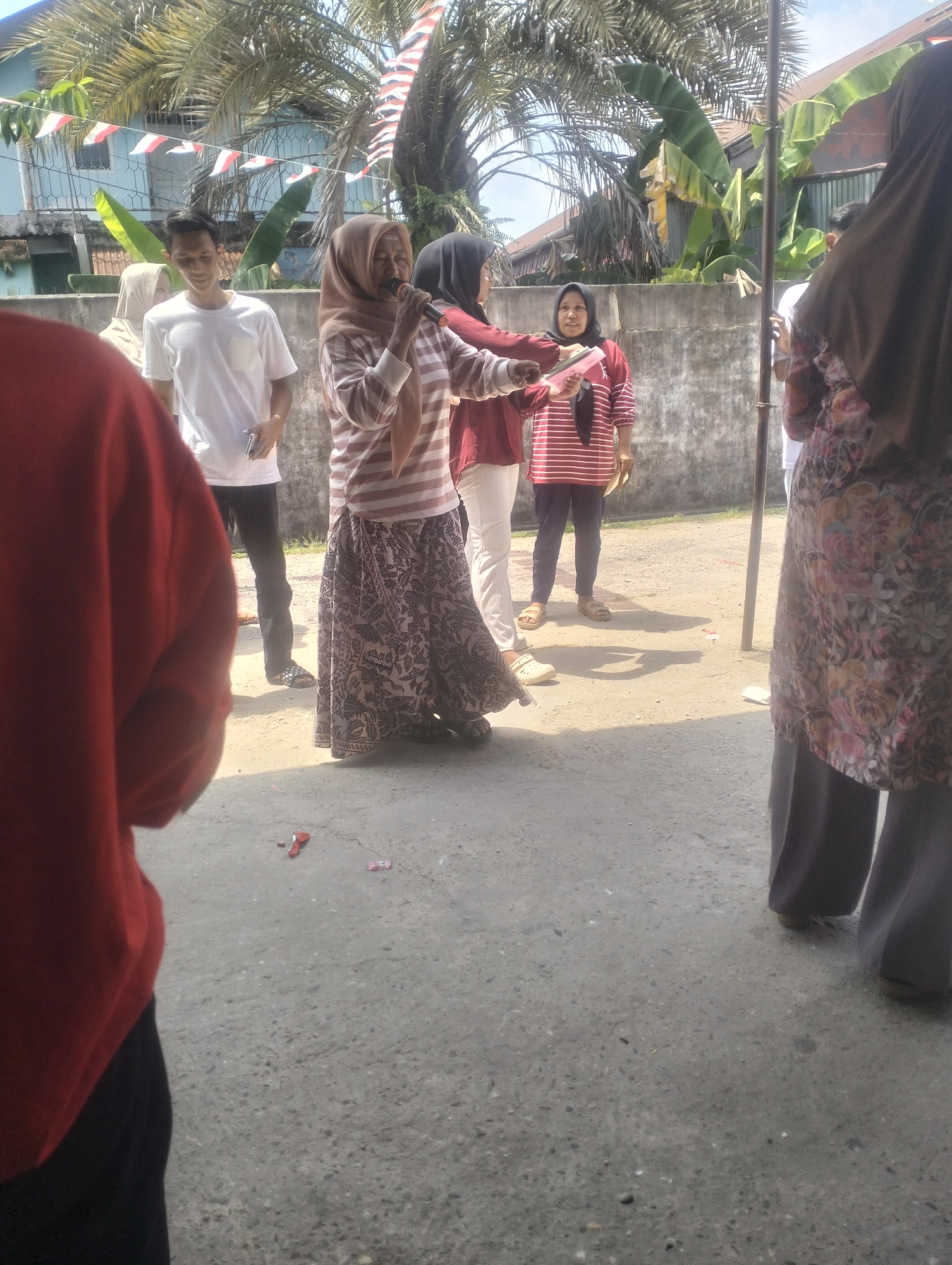 The image depicts an outdoor scene with several individuals. In the foreground, a person wearing a red garment is partially visible. Further back, a woman in a hijab and a striped shirt is holding a microphone and speaking. Beside her, another woman in a red and white striped shirt and dark pants stands. To the left of the woman with the microphone, a man in a white t-shirt and dark pants is visible. Behind the main group, a wall and some foliage, including banana trees and palm trees, are present. The ground appears to be a paved surface. The presence of Indonesian flags suggests a national event or celebration. The overall scene suggests an informal gathering or event taking place outdoors in an urban or semi-urban environment within Dumai City, Indonesia, potentially on a sunny day.