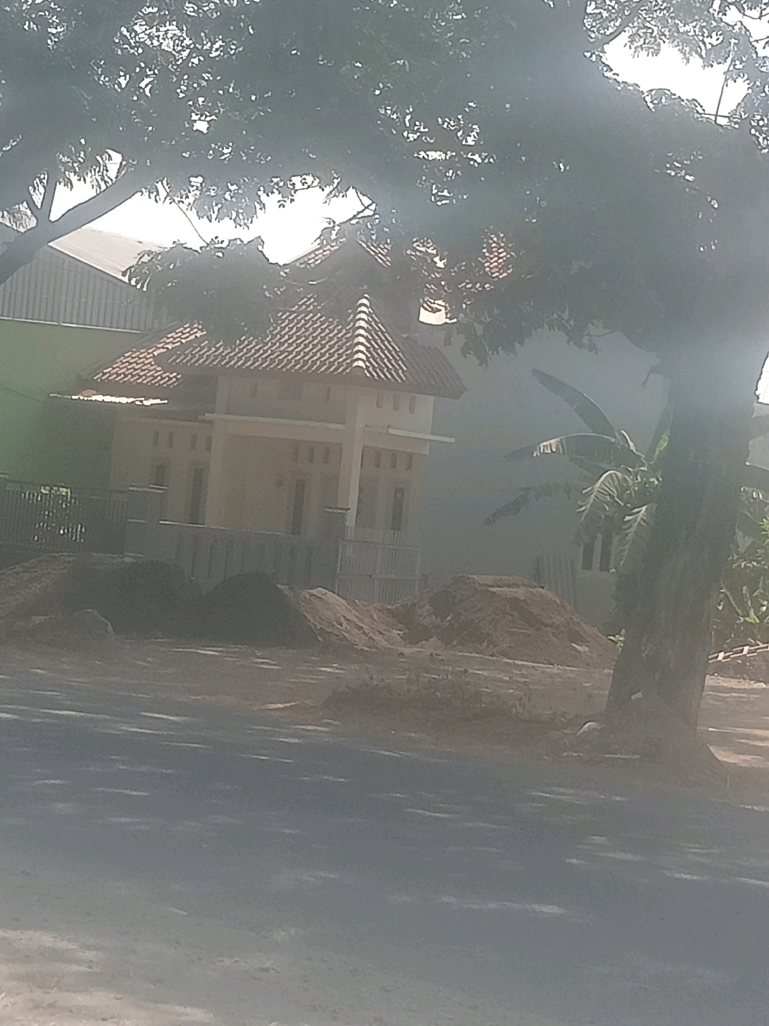 The image depicts a street view in Penusupan, Indonesia, featuring a building under construction. The building, with its characteristic tiled roof, is partially obscured by a large tree in the foreground. Piles of dirt and sand are visible around the construction site, indicating ongoing work. A banana tree can be seen to the right of the building. The overall scene suggests a typical day in a developing area, with elements of nature and construction coexisting.