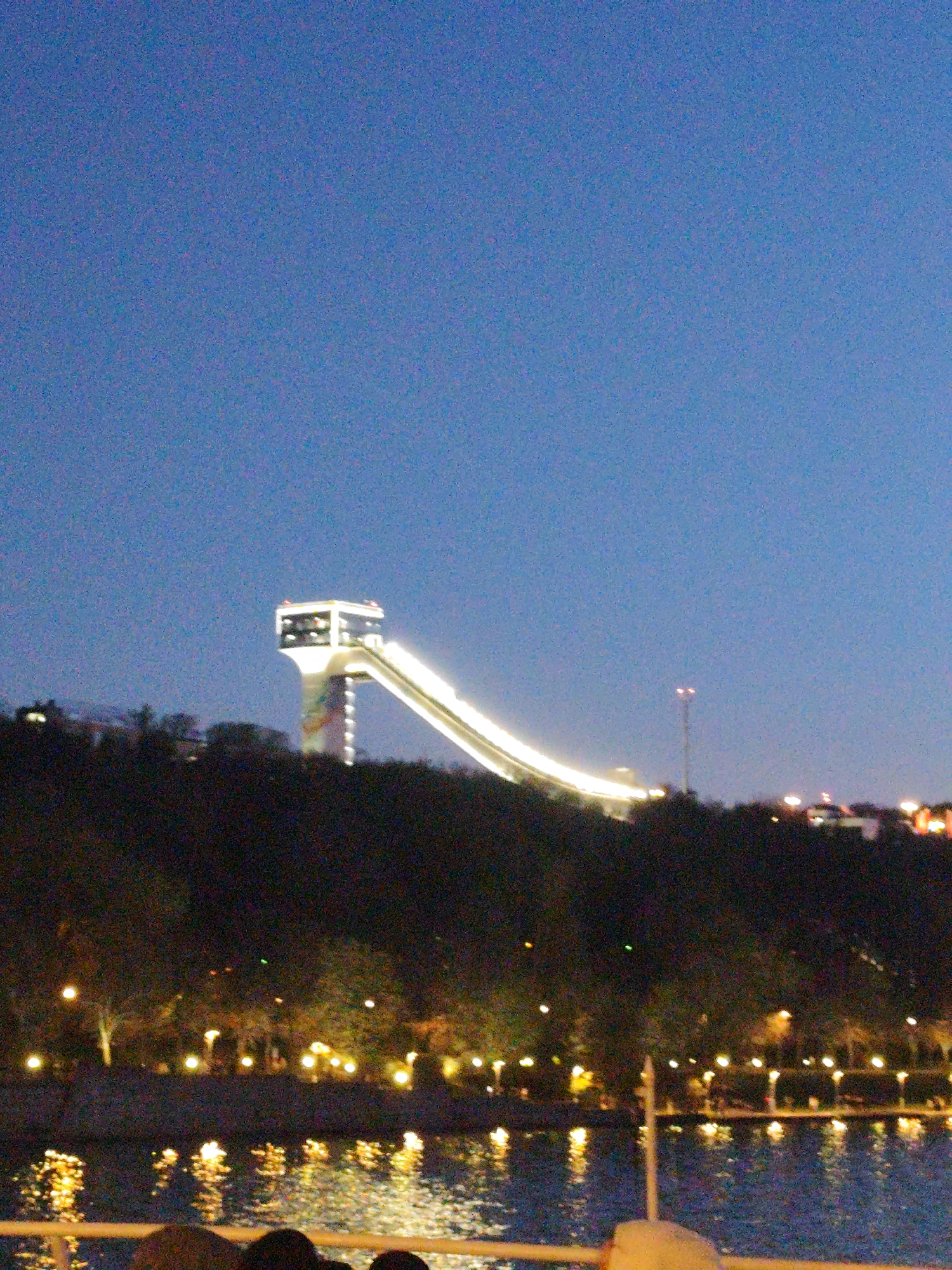 A night scene depicts a cityscape with water in the foreground. Lit trees line the water's edge. Behind the trees rises a dark, treed hill. A large, lighted ski jump structure is situated atop the hill. Lights from buildings are visible on the horizon. The sky is a gradient of blue. Parts of people are visible in the very foreground.