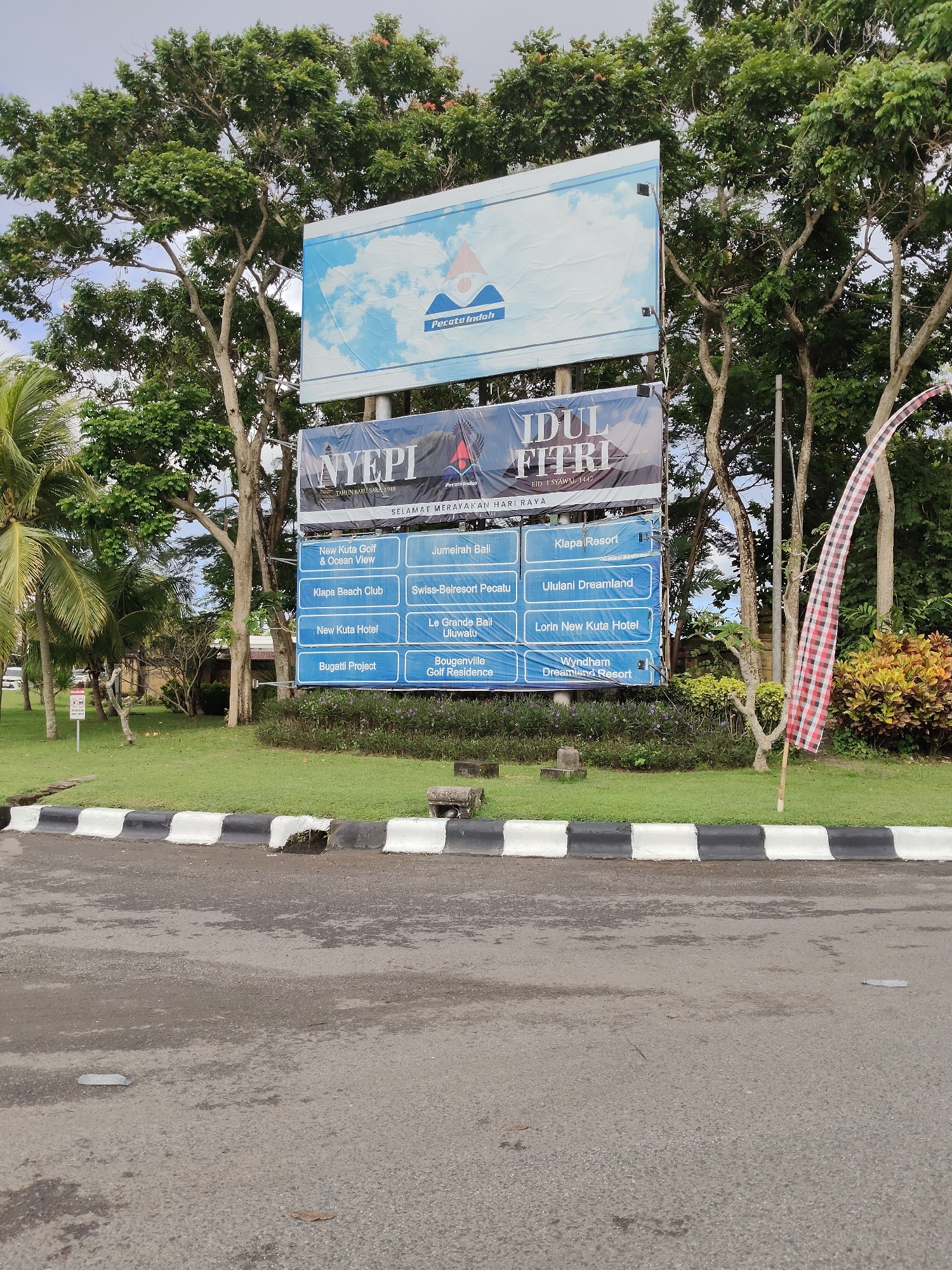 The image shows a large informational sign in Ungasan, Indonesia, surrounded by trees and greenery. The sign is layered with different panels. The top panel features a logo with blue and white imagery. Below that are two smaller signs, one for "Nyepi" (Balinese New Year) and one for "Idul Fitri" (Eid al-Fitr). Beneath those are six panels listing various local resorts and attractions, including New Kuta Golf & Ocean View, Klapa Beach Club, Jumeirah Bali, and Klapa Resort. A black and white striped curb runs along the bottom of the image, separating the sign from an asphalt road. A red and white checkered flag is placed on the right side of the sign. The sky is overcast.