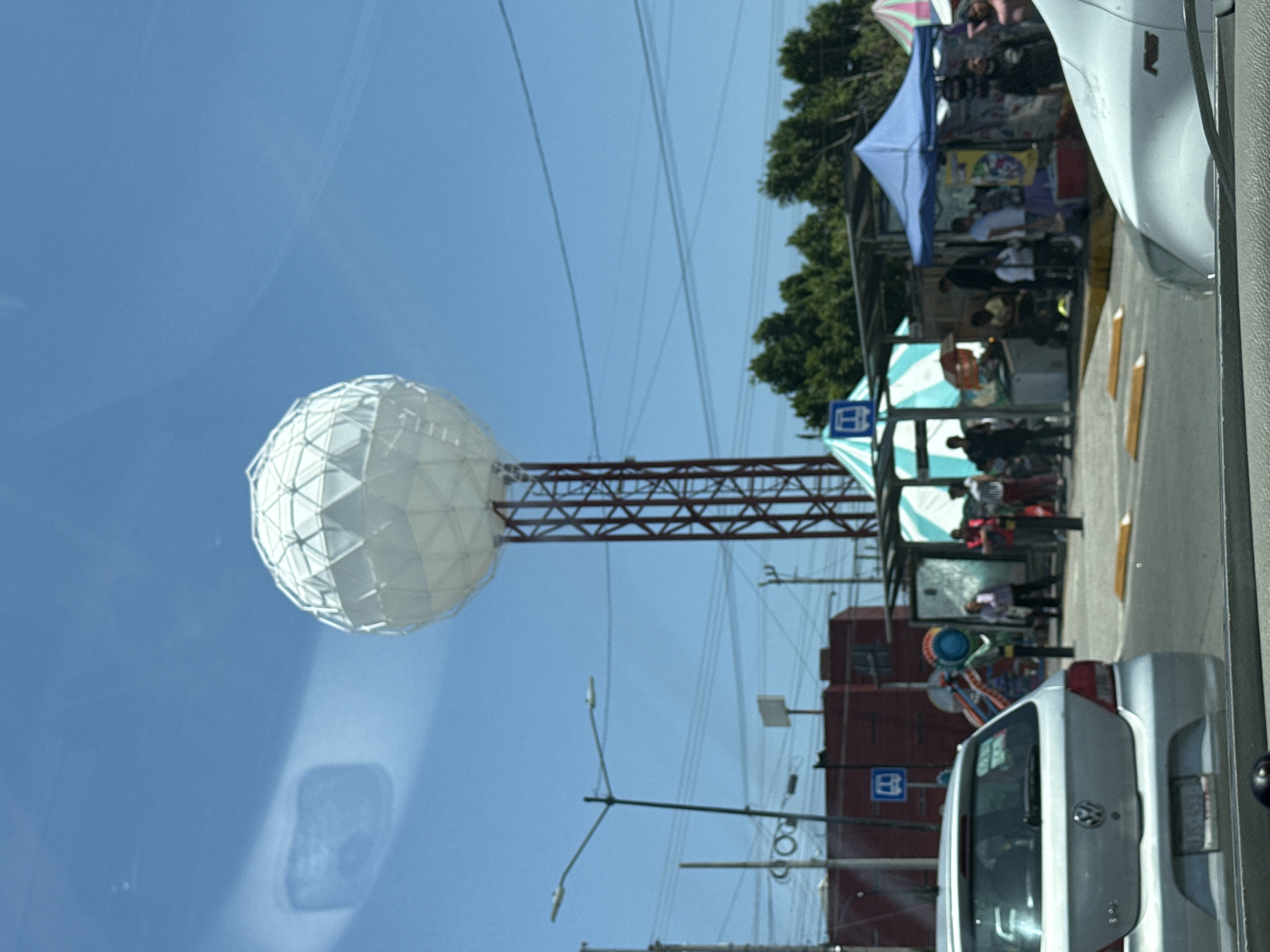 A daytime outdoor scene is depicted under a clear, bright blue sky. On the left, a prominent white geodesic sphere, constructed from geometric panels, is mounted atop a tall, brown metal lattice tower. Numerous thin power lines crisscross the sky above the sphere and extend across the upper portion of the image.
On the right side of the frame, an active street market or fair is in progress. Several colorful vendor stalls are visible, sheltered by awnings in shades of blue, green and white stripes, and pink and white stripes. Multiple people are scattered throughout this area, appearing to be vendors and customers engaging with the stalls. Green foliage from trees can be seen in the background behind the market.
In the foreground on the right, the rear of a silver Volkswagen car is clearly visible, parked or moving on a paved surface with yellow road markings. Another vehicle is partially obscured further down the street. Two blue square signs featuring a white stylized 'B' symbol (likely indicating a bus stop) are visible: one on a pole near the market stalls and another on a reddish-brown building in the background. The overall impression is a bustling urban environment under sunny conditions.