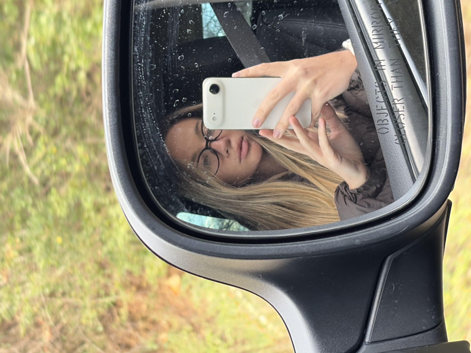 The scene captures a reflection of a female individual in the passenger-side wing mirror of a vehicle, located in an outdoor area of Unknown, Unknown, characterized by blurred green and brown foliage in the background. The individual, who has long blonde hair and wears black-framed glasses, is dressed in a brown jacket and is secured by a seatbelt visible across her chest, indicating she is inside the vehicle. She is holding a light-colored smartphone with both hands, using its camera to photograph her reflection in the mirror. The mirror's surface displays numerous small water droplets or smudges. The text "OBJECTS IN MIRROR ARE CLOSER THAN THEY APPEAR" is visible along the lower edge of the mirror glass.