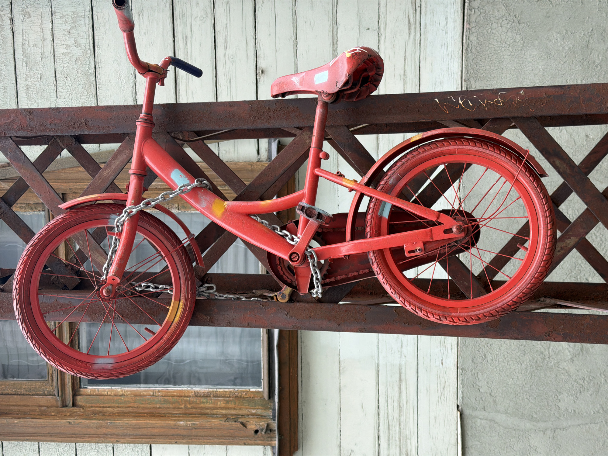 An entirely red-painted children's bicycle is prominently displayed, mounted horizontally on a weathered, rusty metal beam structure. The bicycle, including its frame, handlebars, seat, tires, spokes, pedals, and chain, is uniformly painted in a vibrant red, showing some signs of wear and spray paint marks in yellow and light blue. It is secured to the metal beam by a silver chain wrapped around its front forks and the bicycle's frame.
The metal beam, also reddish-brown and heavily rusted, features a crisscross lattice pattern beneath it, suggesting it might be part of a fence, balcony, or decorative installation. Against the beam, faintly scratched or painted in gold, are letters that appear to spell "WERK" or "WERKS." Further details on the bicycle tires include faint, painted-over white markings, possibly original tire text, with some numbers like "2023" or "2013" barely discernible.
In the background, a light-colored wooden wall, possibly painted white or off-white, shows significant peeling and cracking paint, indicative of age and exposure to elements. Below the metal structure, a portion of an old wooden-framed window with visible glass panes can be seen, reflecting some light. The scene has an aged, rustic, or artistic feel, with the bright red bicycle contrasting against the distressed textures of the metal and wood. The overall impression is that of a decorative street art piece or a unique architectural detail in Riga, Latvia.