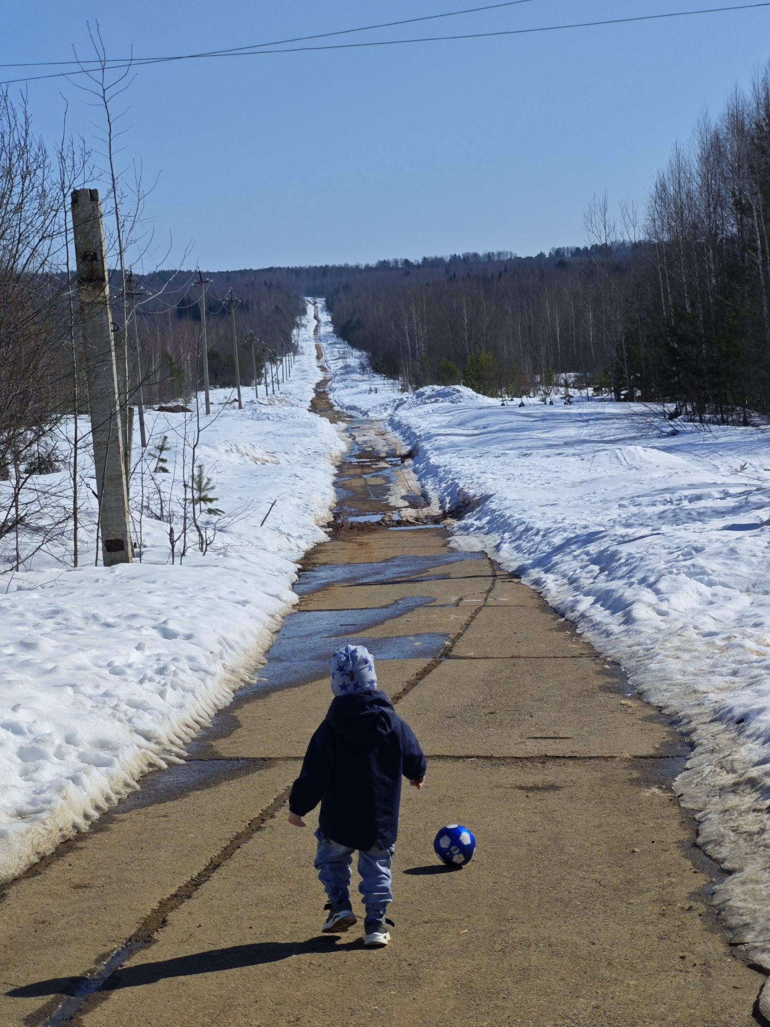 A long, slightly uphill, concrete road bisects a snowy landscape in Марковский, Russia. A small child, seen from the back, wearing a blue jacket, jeans, and a patterned hat, walks away from the viewer down the road, a blue soccer ball beside them. Patches of melting snow create pools of water on the road's surface. The road is lined with snow on both sides, giving way to bare trees and a dense forest in the distance. Several concrete utility poles run alongside the road on the left. The sky is clear and blue.