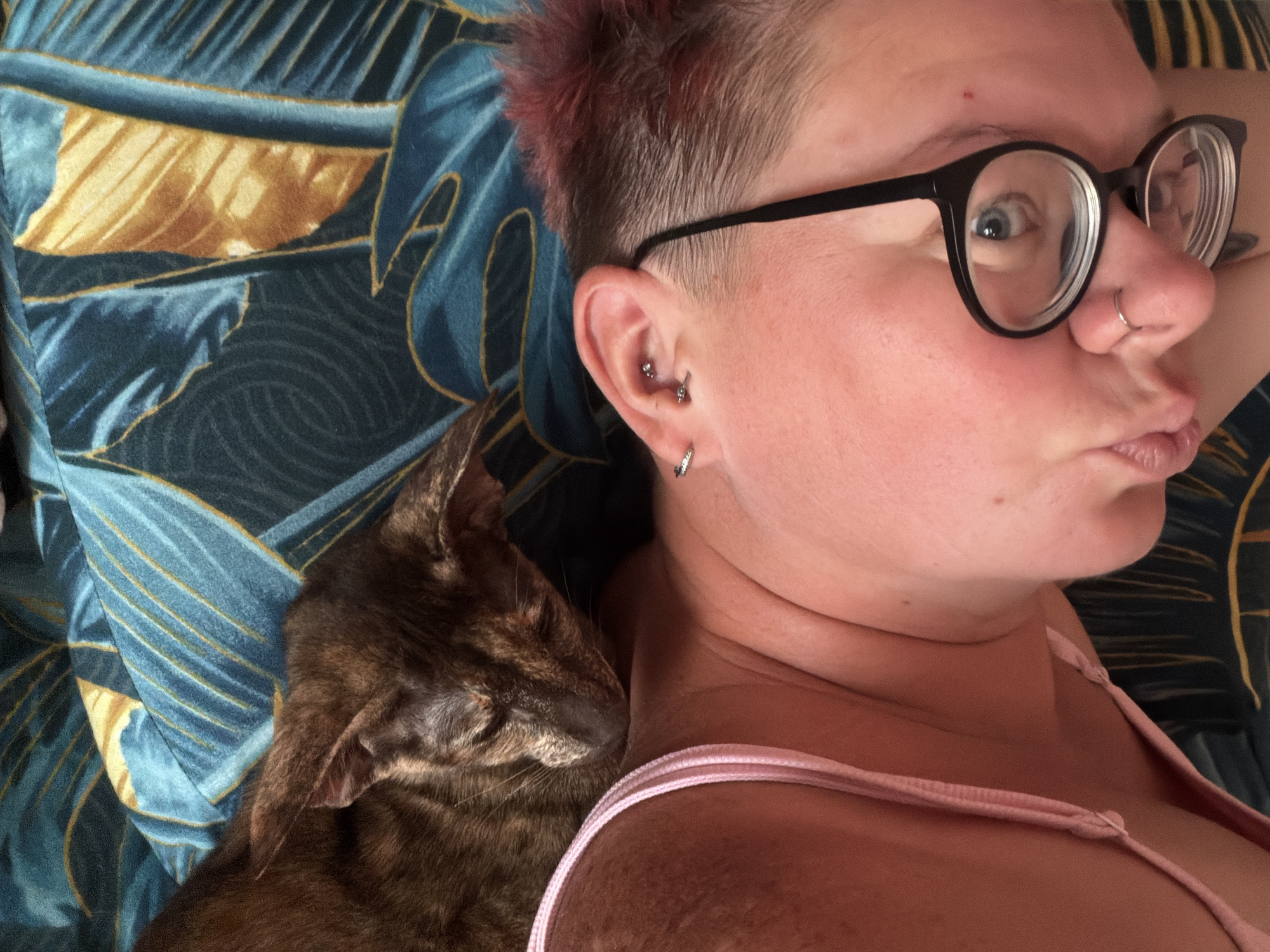 This is a close-up, overhead, first-person perspective photograph of a person lying down with a cat sleeping on their chest. The person has short, reddish hair and is wearing glasses. The cat is a dark tabby with green eyes. The background is a blue and gold tropical print fabric. The image is taken indoors, and there is no indication of the city of Chaikovsky, Russia.