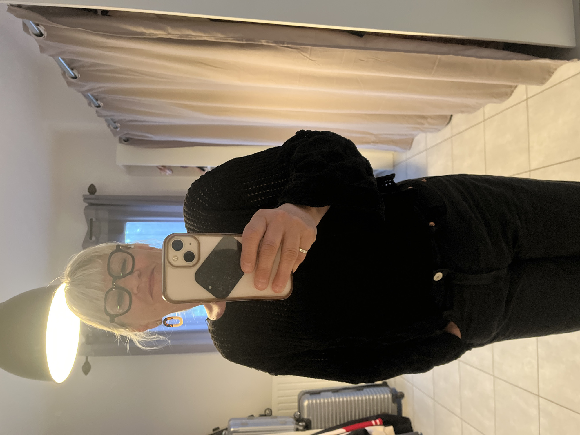 The image is a selfie taken in a room with a light-colored tiled floor. A woman with short blonde hair and glasses is holding a smartphone. She is wearing a black sweater and black jeans, with one hand in her pocket. In the background, there is a curtain, a radiator, and luggage. The city is Chambœuf, France.