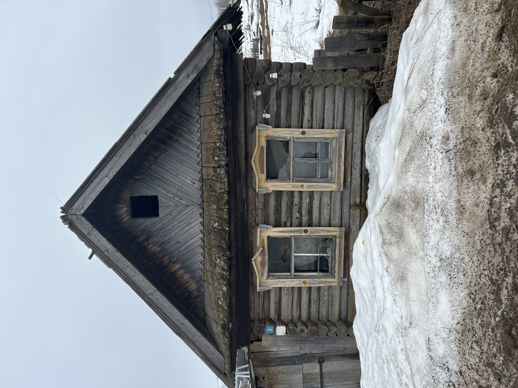 An exterior shot of a dilapidated wooden house in Уральское, Russia. The house has a triangular gable end with a small square window. The lower part of the house has two windows with broken glass panes and peeling yellow frames. The house is constructed from stacked logs. Patches of snow are present around the base of the house and along a fence made of wooden planks.