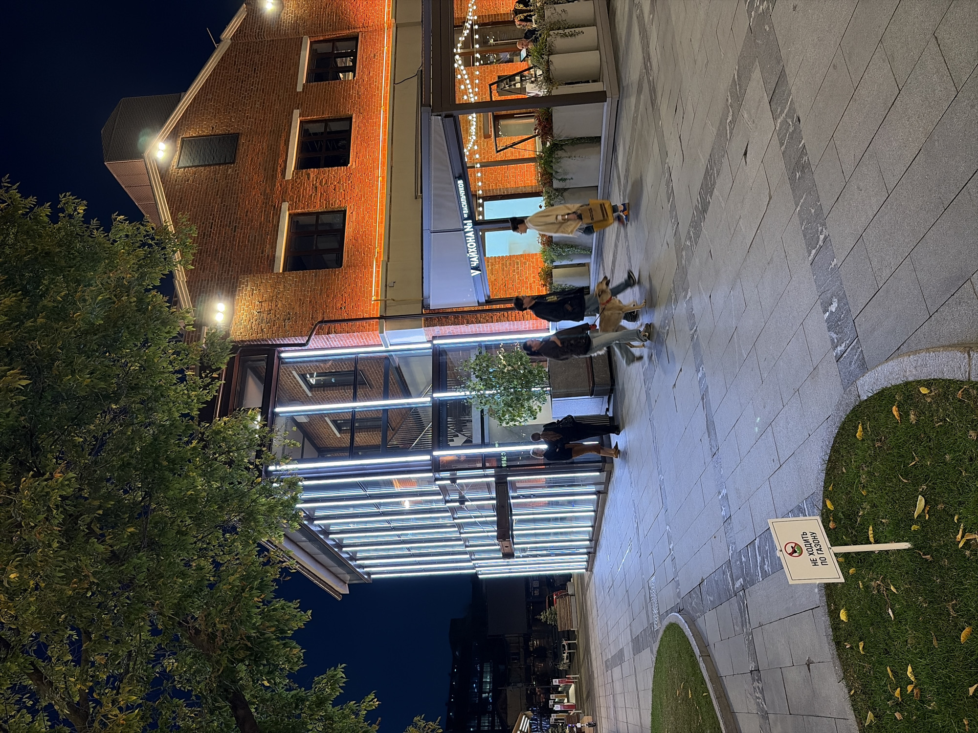 The image depicts an outdoor scene at night, likely in Moscow, Russia, showcasing a contemporary architectural complex illuminated by various lights. A two-story brick building with orange-tinted lighting forms the backdrop, featuring large windows and an upper section with a pitched roof and external lights. Adjacent to it is a modern section of the building with a glass facade, vertically illuminated by numerous cool-toned LED lights, creating a striking visual contrast.
In front of these buildings, a paved walkway with decorative stone inlays stretches across the scene. Several people are visible walking on this pathway. On the left side of the image, near the glass-fronted building, two individuals are standing. Further to the right, a group of three people, accompanied by a dog on a leash, are walking. One person in this group is carrying a yellow tote bag.
An outdoor seating area of a restaurant or cafe is visible to the right, adorned with string lights and planters. In the foreground, a patch of green grass has a white sign with a red circle and a line through a dog icon, along with Russian text that translates to "Do not walk on the lawn." Fallen yellow leaves are scattered on this grassy area and the surrounding pavement. The overall atmosphere is urban and modern, illuminated by artificial light against a dark night sky.