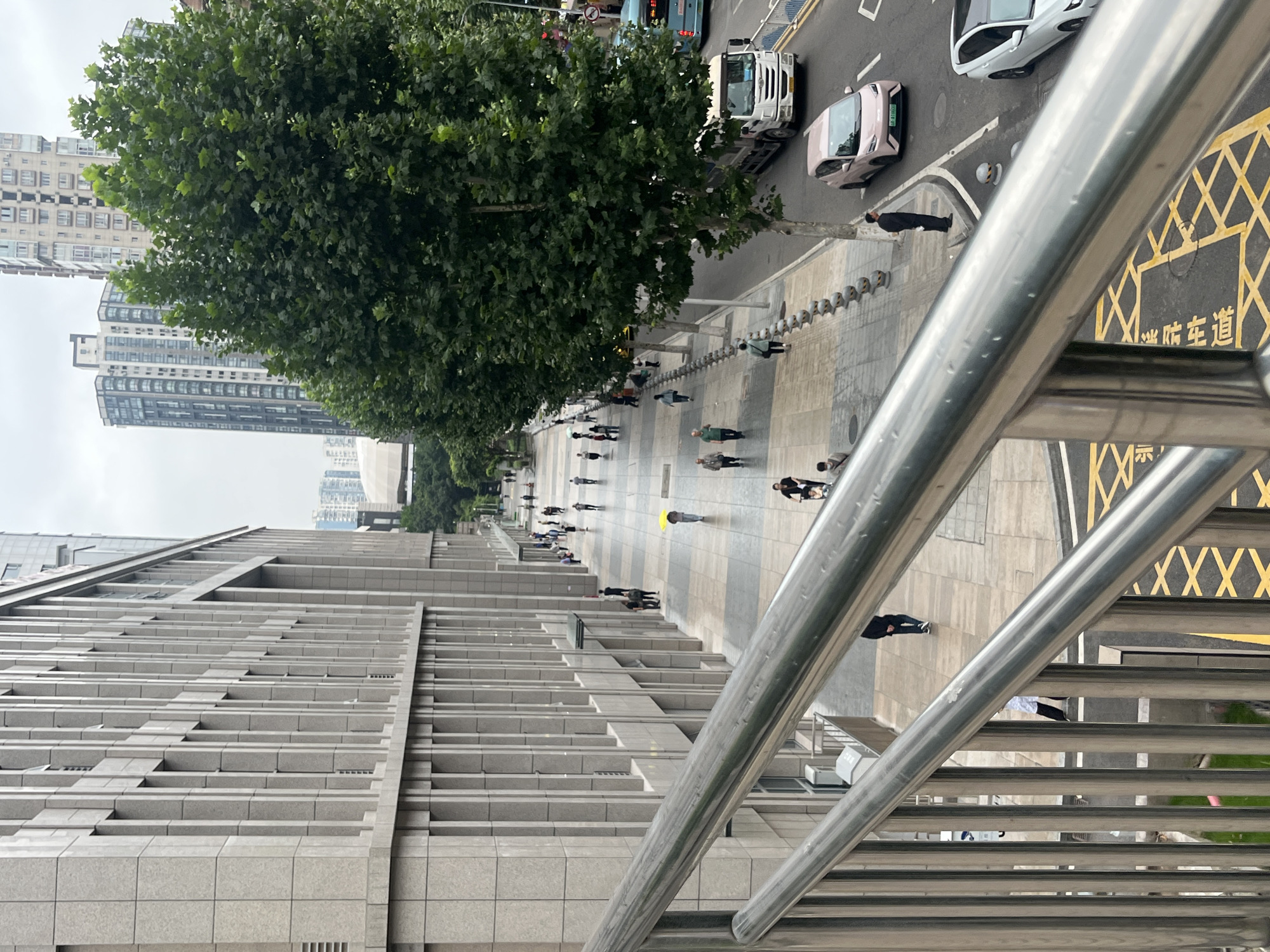The image captures a street scene in Yuzhong District, China, viewed from a pedestrian bridge with prominent silver railings in the foreground. A large, modern building with a light-colored, textured facade and vertical window elements dominates the left side of the frame. The sidewalk adjacent to the building is paved with light-colored tiles and features a row of grey, spherical bollards.
Several people are walking along the sidewalk, some individually and some in small groups. One person is holding a bright yellow umbrella. A few trees with full green canopies are planted along the edge of the sidewalk, separating it from the street.
The street itself is paved with asphalt and shows lane markings. Traffic is present, including a white truck, a light pink car, and a white car. A blue bus is visible further down the street. The background features tall, multi-story buildings, some with modern architectural designs. The sky is overcast, suggesting a cloudy day. Yellow painted lines on the road indicate specific traffic or parking regulations.