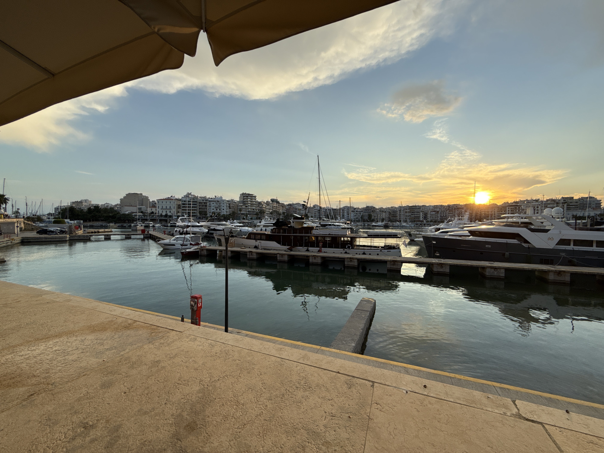 The image shows a harbor scene in Piraeus, Greece at sunset. Multiple yachts and boats are docked in the water, reflecting the sky and surrounding buildings. A concrete pier and walkway are visible in the foreground. A red fire safety box stands on the pier. Buildings line the harbor, illuminated by the setting sun. The sky is a mix of blue and yellow, with scattered clouds. An awning is visible in the top-left corner.