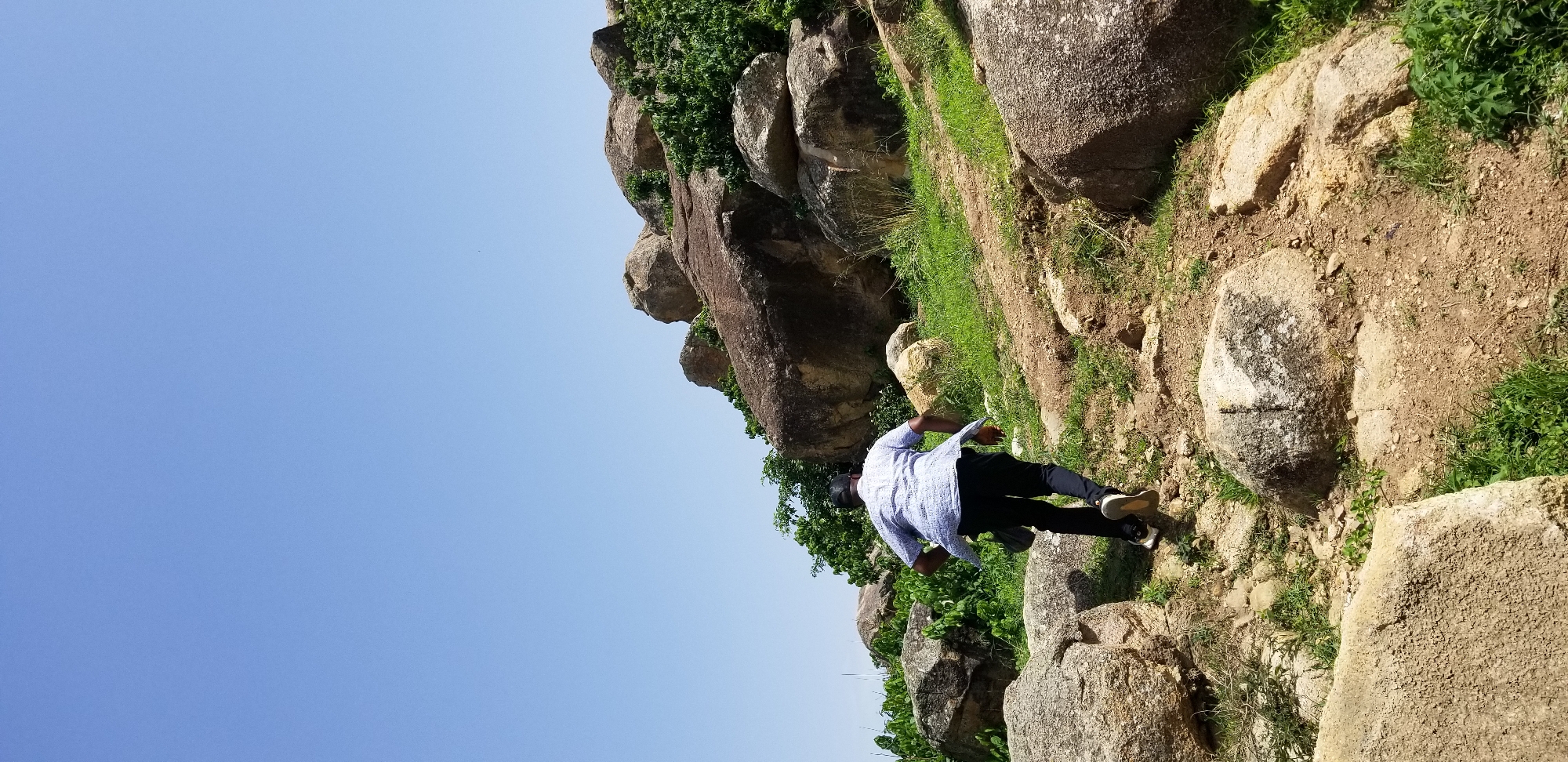 The media file depicts a person, viewed from behind, on a rocky, grassy incline. The individual is wearing a light blue patterned shirt and dark pants, with sneakers on their feet. They appear to be walking or posing on the uneven terrain, which is covered with a mix of dirt, rocks, and green vegetation. Large boulders are scattered throughout the scene, some partially overgrown with grass and small plants. The sky above is a clear, bright blue, suggesting a daytime setting with fair weather. The overall impression is of an outdoor adventure or exploration in a natural, rocky landscape. No text is visible in the image.
