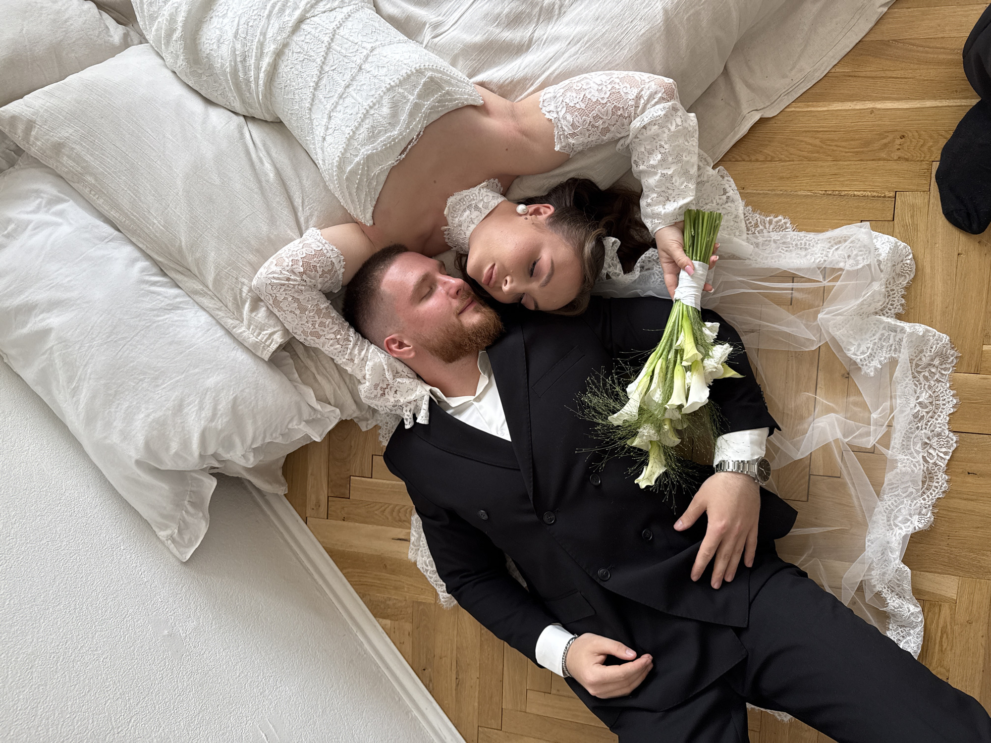 Two individuals, a male and a female, are depicted from an overhead perspective, lying on their backs on a light brown herringbone parquet floor and white bedding in Perm, Russia. The male, positioned on the left, has a beard and short dark hair; he is wearing a black double-breasted suit jacket over a white collared shirt, and black trousers. A silver watch is on his left wrist and a silver bracelet on his right. His eyes are closed, and his head is turned towards the female. The female, positioned on the right, has dark hair styled and closed eyes; she is wearing a white textured lace dress with long sleeves, a white lace choker, and a pearl earring on her visible left ear. Her head is also turned towards the male. She holds a bouquet of white calla lilies and green foliage, wrapped in white ribbon, with her right hand. White bedding, including pillows with varied textures and ruffles, is arranged beneath the female and extends behind both individuals. A sheer white veil with lace trim is draped across the wooden floor near the male's legs and the female's arm. A textured white wall with a white baseboard runs along the upper left edge of the scene. A small black fabric item is visible in the bottom right corner of the frame.