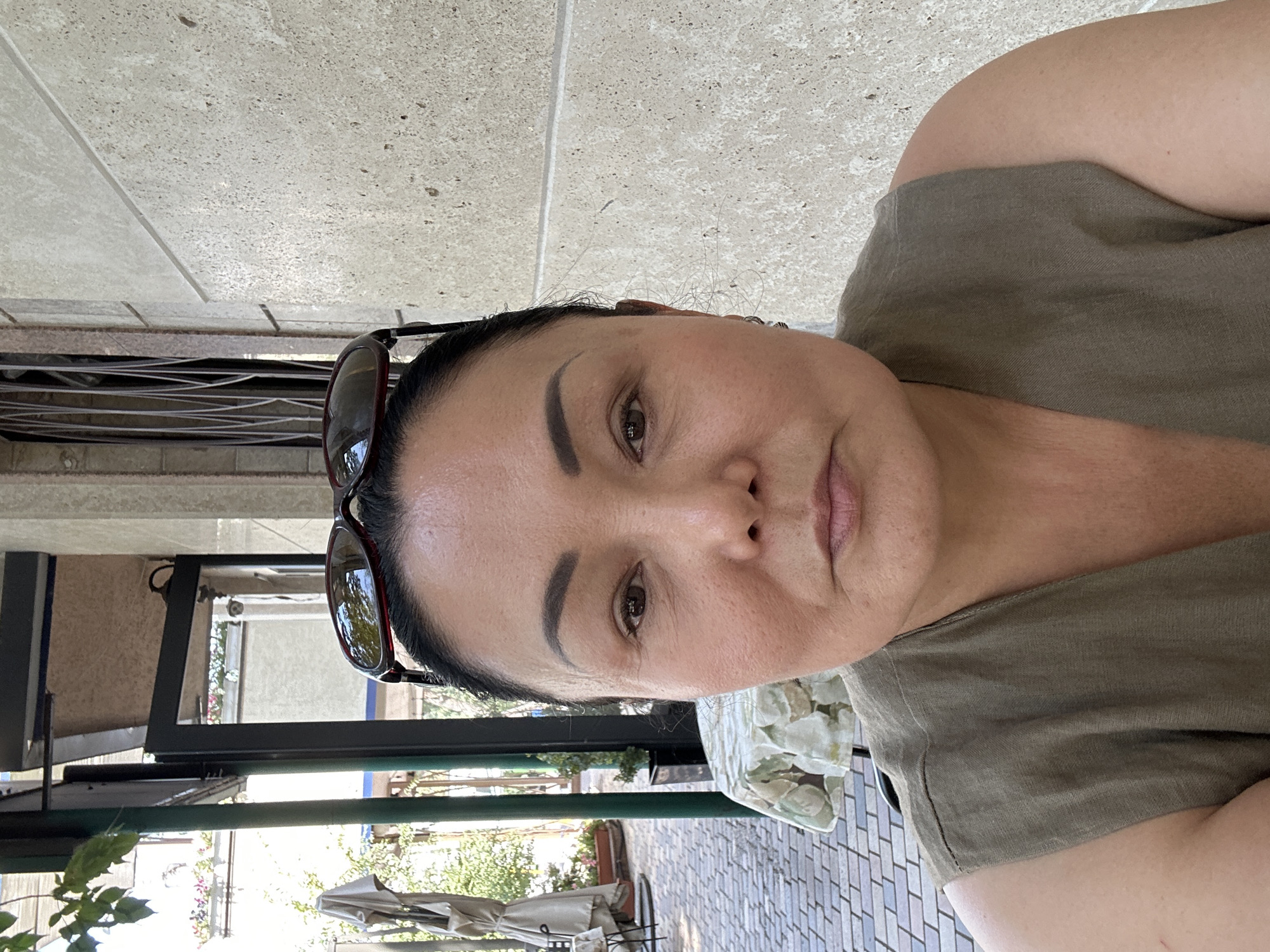 This is a close-up, eye-level, frontal portrait of an East Asian woman in her late 40s or early 50s. She has dark hair pulled back, a fair complexion, and is wearing sunglasses on top of her head. Her makeup is subtle, with emphasis on her eyebrows and a touch of blush. She is wearing a simple, olive-green, sleeveless top.
The background suggests an outdoor seating area, possibly a cafe or restaurant. To the left, there is a table with a patterned tablecloth, and beyond that, a brick-paved walkway and some greenery. To the right, a textured concrete wall is visible. The lighting is natural and somewhat diffused, indicating daytime. The woman's expression is neutral, with her mouth closed. The image appears to have been taken with a smartphone due to the close framing and the selfie-like composition.
This scene could be located in the city of Almaty, Kazakhstan, given the architectural and atmospheric elements, particularly the style of the outdoor seating area and the building materials, which are consistent with what might be found in Almaty.