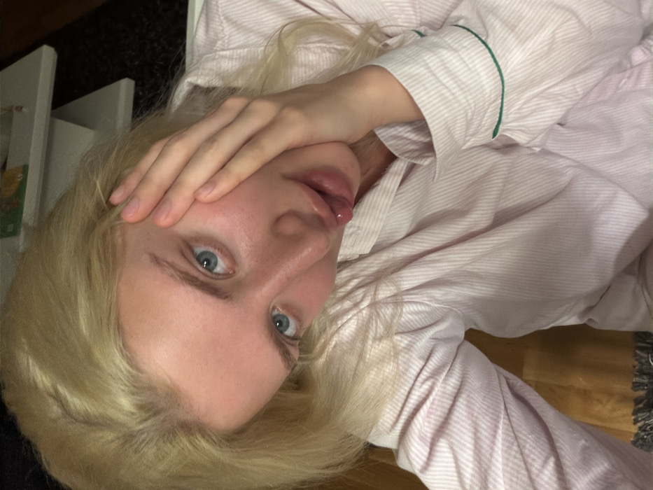 This image captures a close-up, eye-level portrait of a single individual in Budva, Montenegro. The subject is an adult female with blonde hair and blue eyes, presenting a fair complexion with a subtle facial flush. Her head is tilted slightly to the right, and her direct gaze is fixed on the camera. Her left hand is positioned with fingers spread against her left cheek and jawline. Her lips are parted, with the tip of her tongue slightly exposed. She is wearing a light pink and white striped collared shirt, consistent with sleepwear, featuring thin green piping on the cuffs. The background is out of focus and dimly lit, revealing a partial view of a white, multi-tiered shelving unit to the upper right. A segment of light wooden flooring is visible below the subject's left arm, adjacent to a dark, textured floor covering in the lower left.