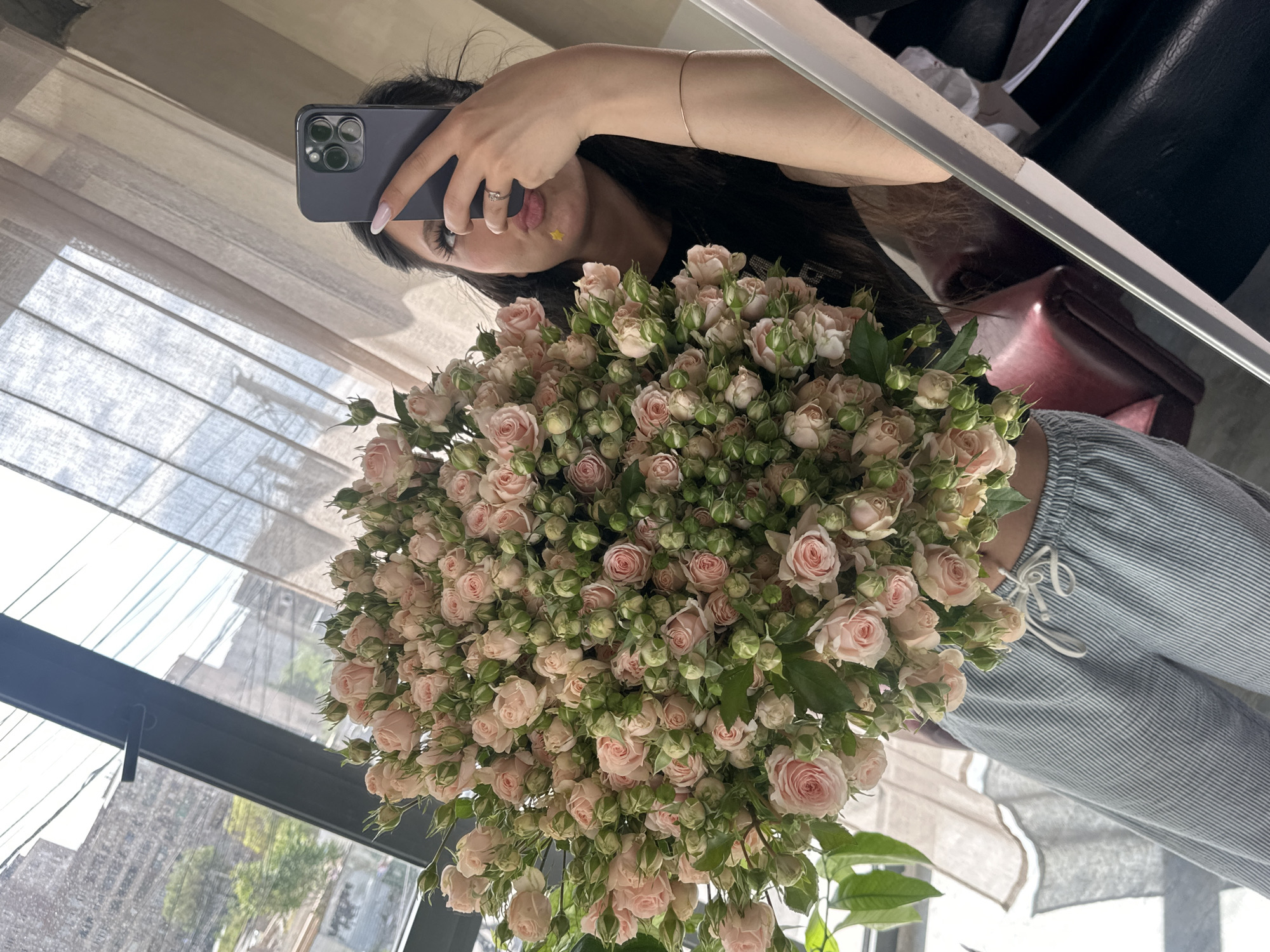 The image, rotated 90 degrees counter-clockwise, depicts a person taking a mirror selfie while holding a large bouquet of light pink spray roses. The individual's face is partially visible, mostly obscured by a dark-colored smartphone (likely an iPhone given its camera setup) held in their right hand. Notable features include a dark eye, nose, and lips, along with long dark hair, manicured nails, a ring on one finger, and a thin bracelet on the wrist. A small yellow star sticker is visible on the lower lip/chin area. The person is wearing light-colored, striped pants with a white drawstring.
The prominent bouquet is dense with numerous light pink roses in various stages of bloom, alongside many green buds and green leaves.
The background, reflected in the mirror, shows an indoor setting with a window and light-colored blinds or curtains. Through the window, an outdoor scene with buildings and some greenery (likely trees) is visible, suggesting it is daytime with clear weather. A dark, possibly leather, object is also reflected in a small section of the mirror. The location context for this scene is Zovuni, Armenia. No visible text is present in the image.