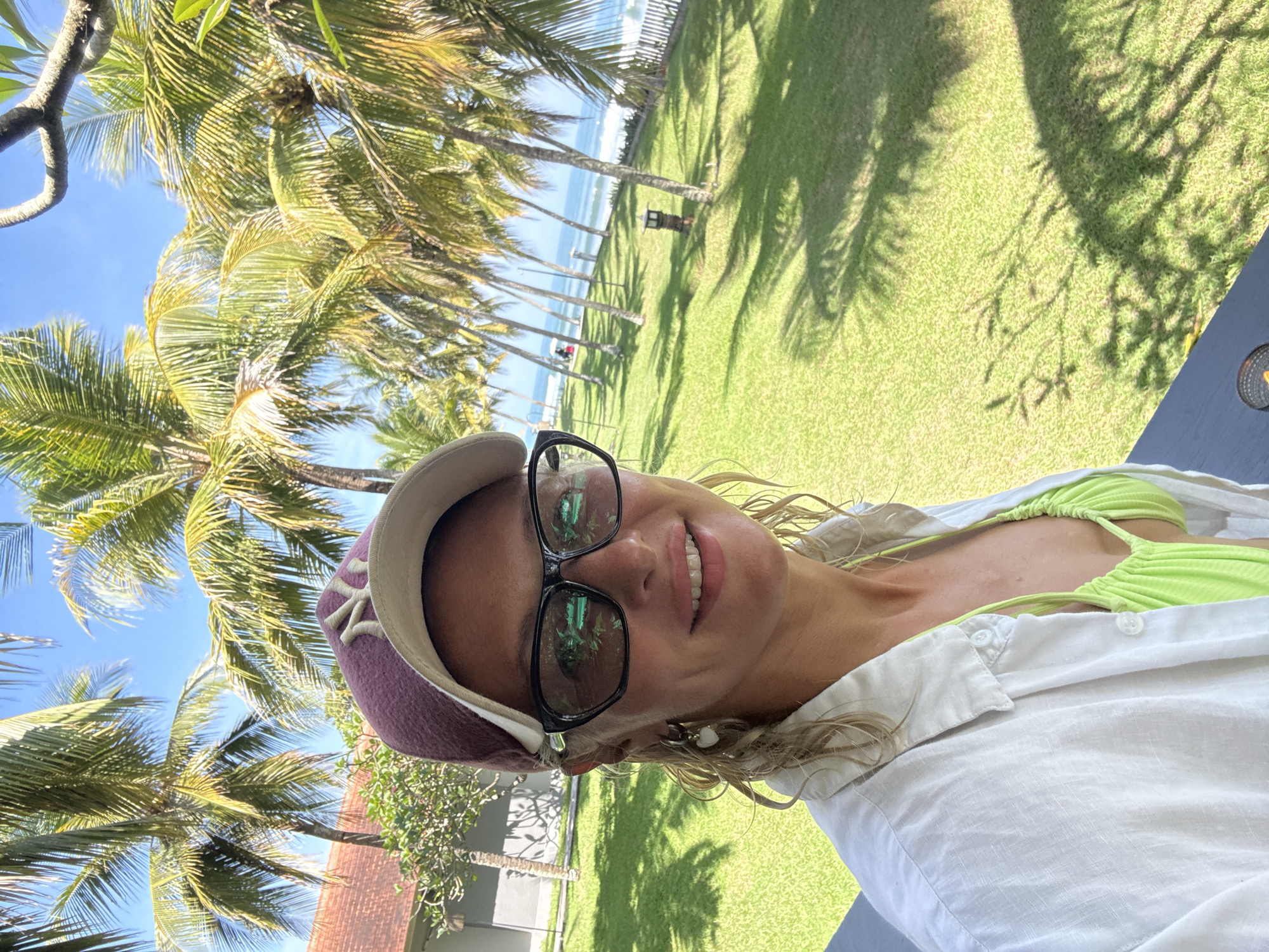 A waist-up shot depicts a smiling woman with blond hair, wearing glasses and a maroon baseball cap, along with a white linen shirt over a lime green bikini top. The background includes a lush green lawn with palm trees that stretches out to a beach and the ocean under a bright blue sky. The location is Unknown, Unknown.