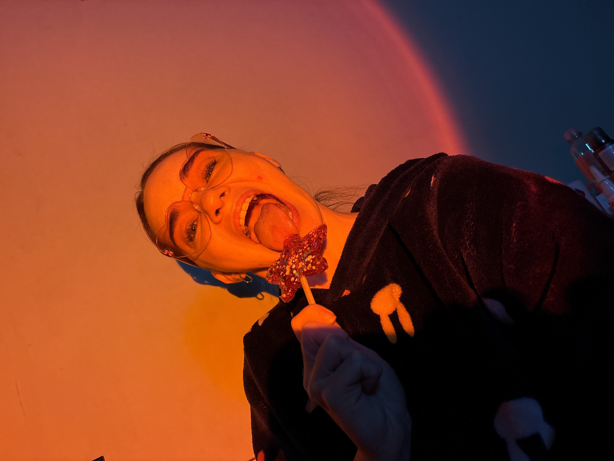 A woman wearing glasses and a dark hooded garment with bunny designs is holding a star-shaped lollipop to her tongue. The scene is lit with orange hues from the left side, which transitions to a blue tone on the right, with a curved line between the colours. A few bottles are visible on the far right. The location is in the city of Unknown, Russia.