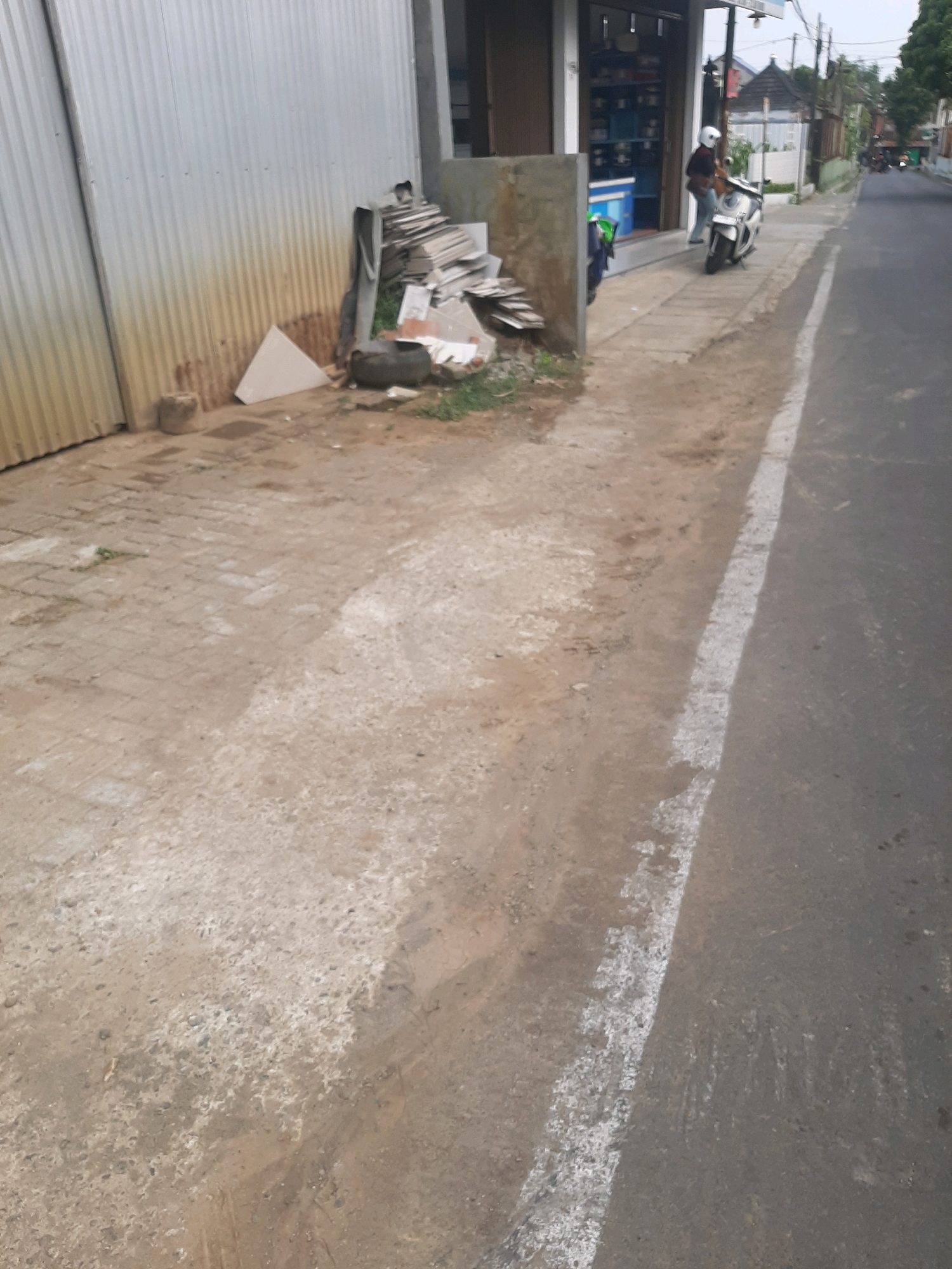 The image depicts a street scene in Purwanegara, Indonesia. A person wearing a white helmet is seated on a white scooter parked on a concrete sidewalk. To the left of the scooter, there is a building with a metal corrugated wall and a storefront. Piles of construction debris, including what appear to be tiles and some fabric, are stacked near the building. The sidewalk leads to a paved road with a white line marking its edge. Further down the road, other buildings and trees are visible, suggesting a residential or commercial area. The overall scene is of a typical street in a populated area, with evidence of ongoing construction or renovation.
