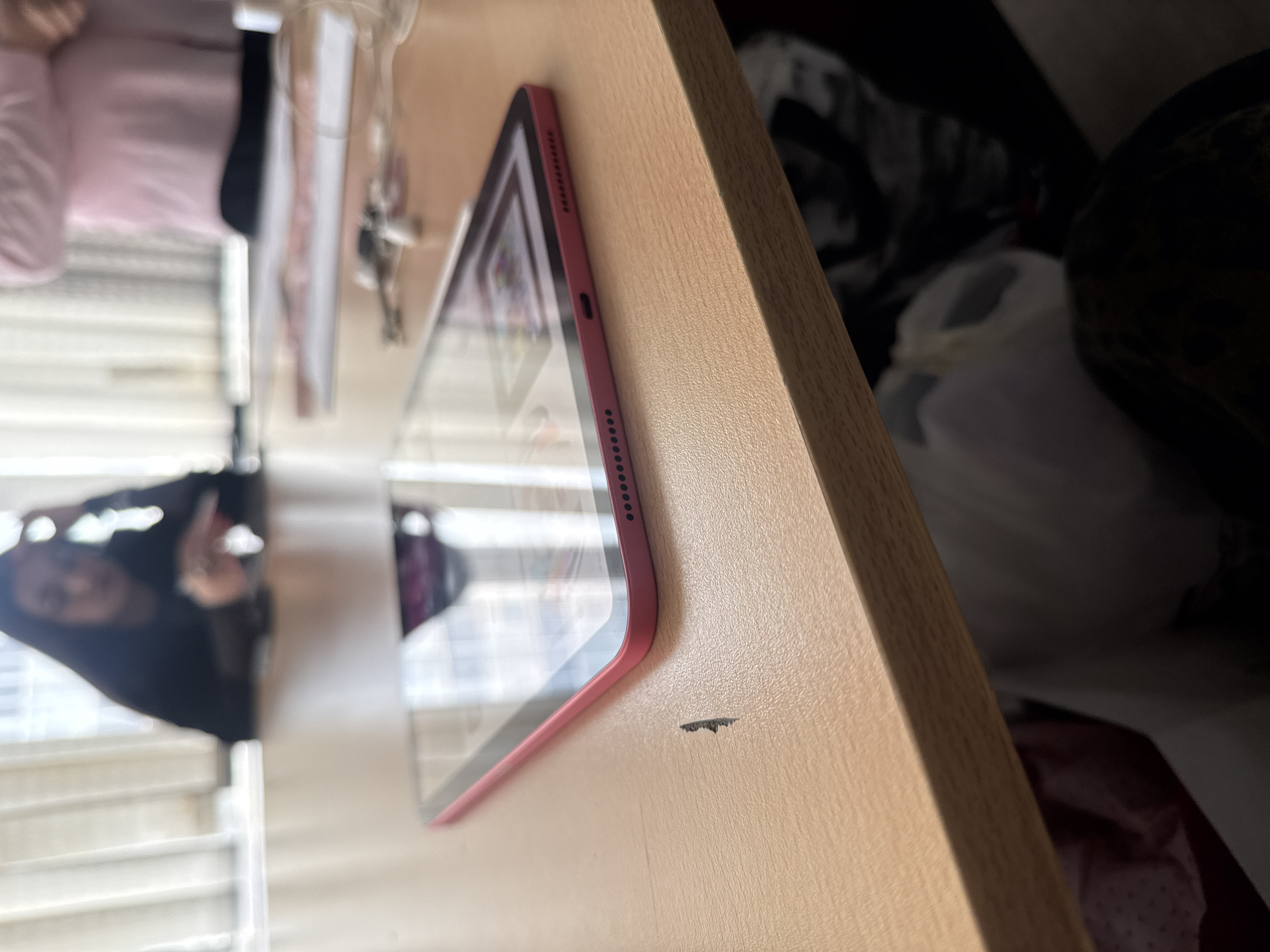 The image shows a pink tablet lying on a light brown wooden surface. The edge of the tablet is visible, showing a series of small holes, likely speaker vents, and a small port. A small dark imperfection is present on the wooden surface near the tablet. The tablet's screen reflects a room with a window, blinds, and a person. A cluttered area with various objects is seen blurred in the background. The precise location in Yerevan, Armenia, cannot be determined from this image.