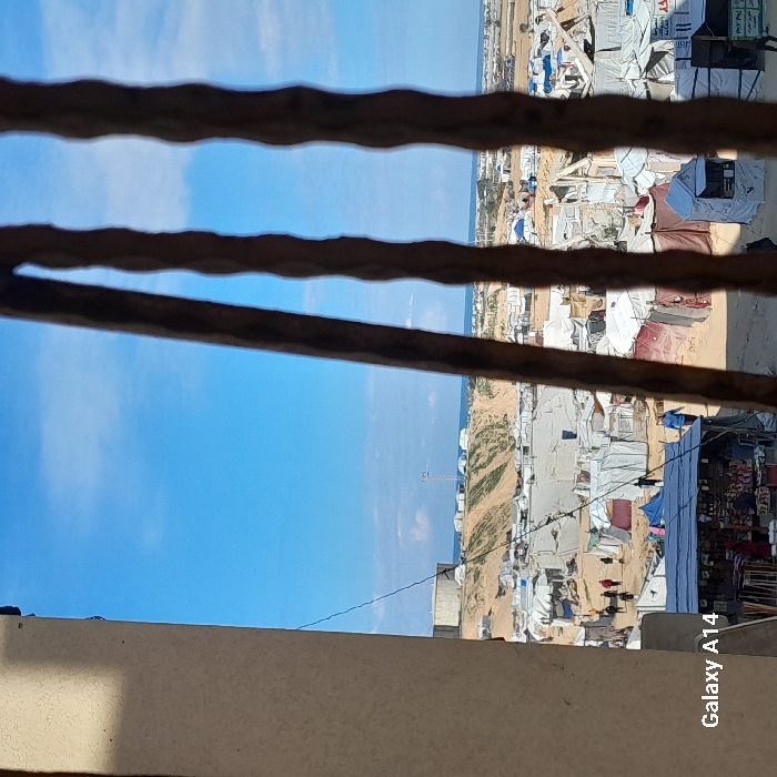 The image displays a panoramic view of a densely populated temporary settlement or refugee camp situated in Khan Yunis, Palestinian Territory. The scene is observed through three vertical, dark-colored bars, likely from a window or gate, which partially obscure the right side of the frame. A light-colored building wall forms the left border.
The midground is dominated by hundreds of tightly packed makeshift tents and shelters, predominantly white and light-colored, constructed from tarpaulins and fabrics. These structures are erected on sandy ground, with visible dirt paths crisscrossing between them. Multiple individuals are seen walking throughout the camp, dressed in civilian attire. In the lower-left portion of the camp, a market stall, covered by an awning, displays various goods, with people present nearby. Some tents in the lower right display partial signage.
In the background, the terrain consists of sandy hills and dunes, upon which additional temporary structures and tents are visible. A large, cylindrical water tank is prominent on a hill in the upper-left. A tall communication tower or mast stands on another distant hill. Beyond these hills, a distinct blue horizon marks the meeting of a clear sky with a body of water, consistent with a coastline and the Mediterranean Sea, with distant coastal development discernible. The sky above is clear with scattered, wispy clouds.