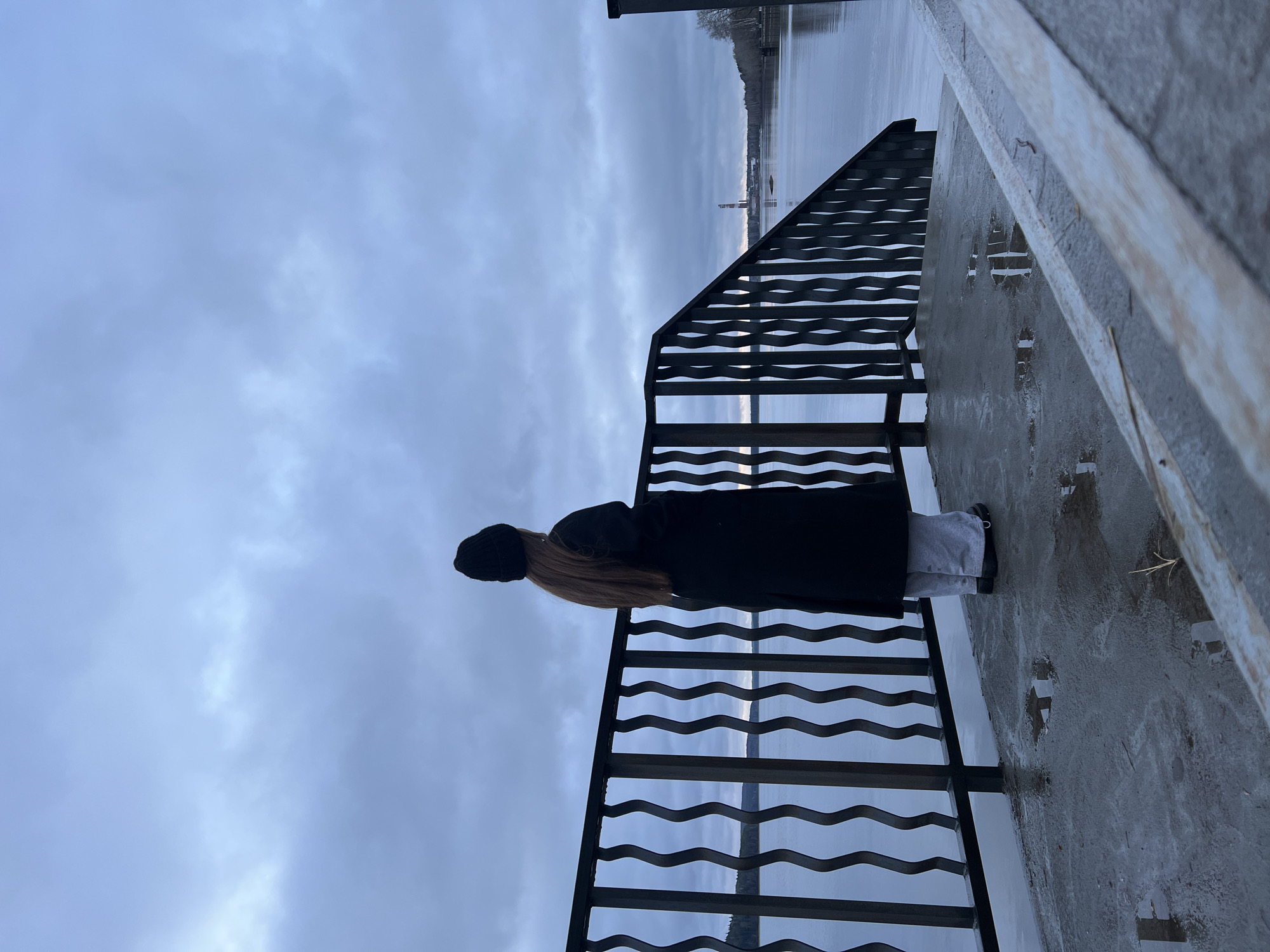 A woman in a black coat and black beanie stands at a wavy, black metal railing, looking out toward a body of water. The railing is situated on a concrete platform that appears wet. The sky is overcast. The location is Chaikovsky, Russia.