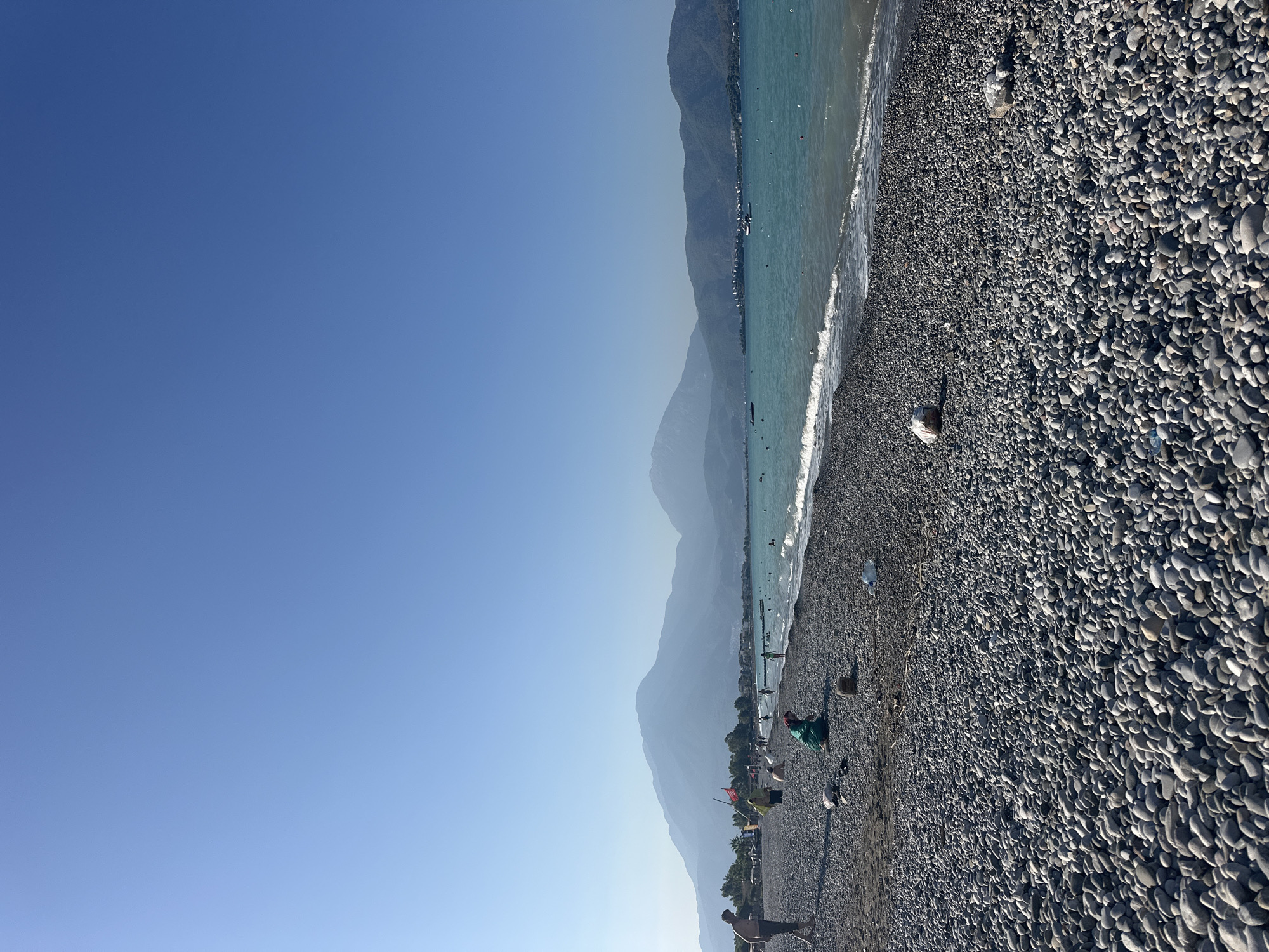The image depicts a wide, pebble-covered beach in Fethiye, Turkey, stretching towards a calm, turquoise sea under a clear blue sky. In the distance, lush green mountains form a picturesque backdrop. Several people are visible on the beach and in the water, enjoying a sunny day. Some are walking along the shoreline, while others are seated or playing in the shallow waters. A few boats are scattered across the sea, suggesting a leisurely atmosphere. The foreground is dominated by the smooth, grey pebbles that characterize the beach. The scene captures a typical summer day at a coastal resort town.